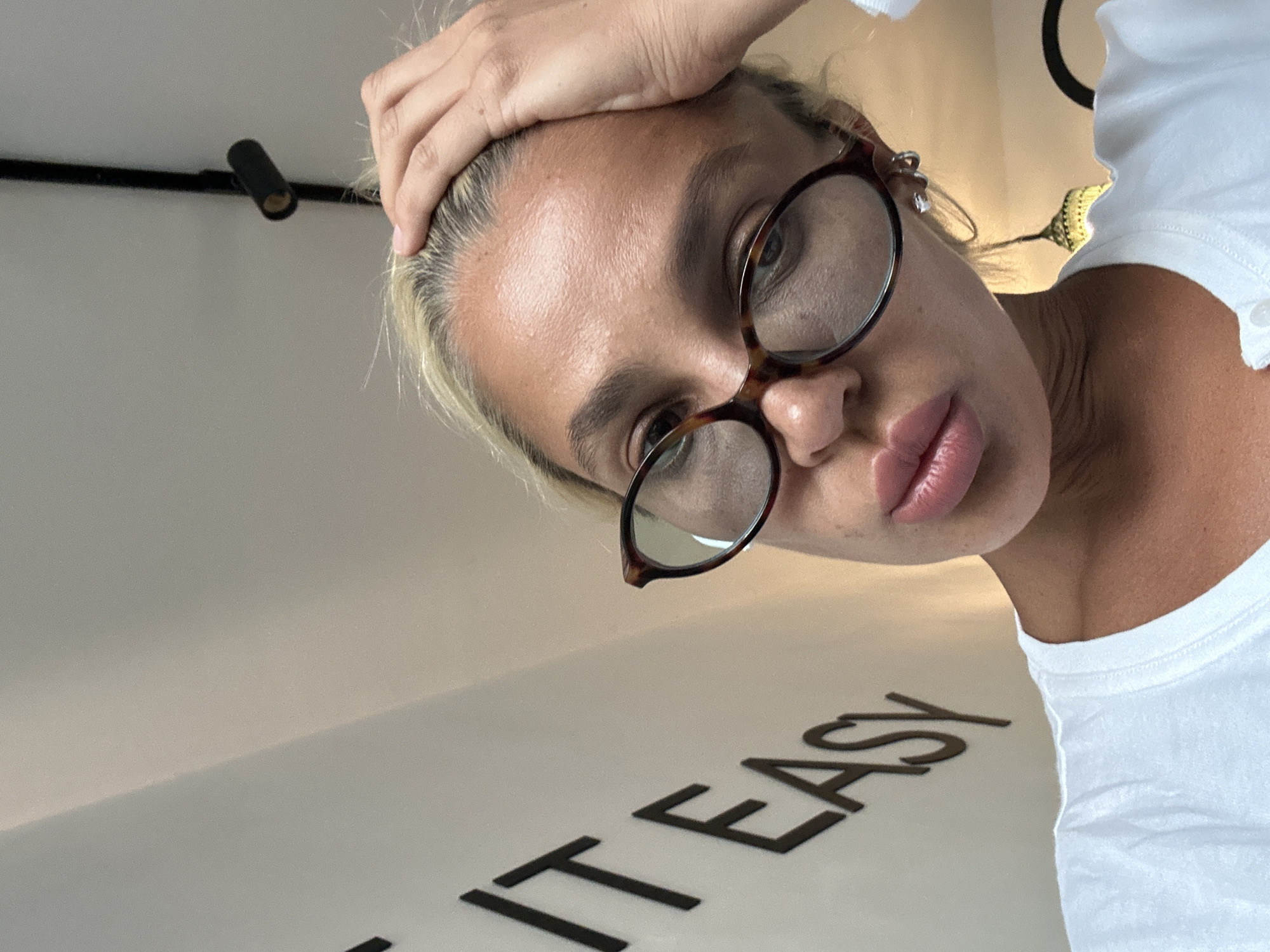 In this close-up, low-angle selfie taken from a slightly elevated perspective, a woman with blonde hair is shown resting her right hand on her forehead. She is wearing large, tortoiseshell-framed glasses and a white t-shirt. Her lips are slightly parted, and she appears to be looking slightly upwards. In the background, on a white wall, the black text "IT EASY" is visible. The lighting suggests an indoor environment, possibly a room or studio, with a decorative light fixture visible in the upper right portion of the frame. There is no indication of the city of Kuta Selatan, Indonesia, within the image itself.