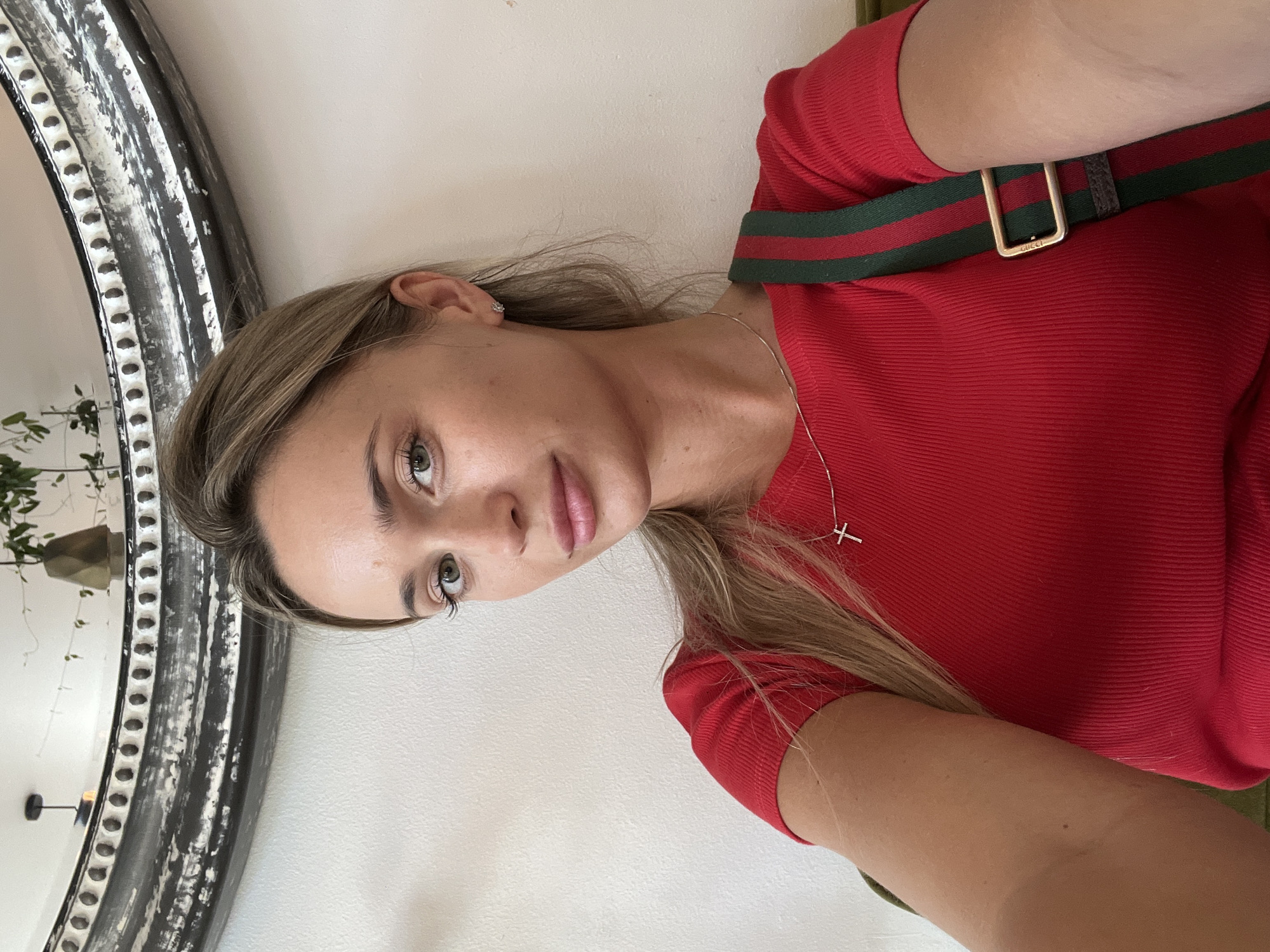 A woman with blonde hair pulled back in a ponytail is looking at the camera in a close-up shot. She is wearing a red ribbed t-shirt and a necklace with a silver cross pendant. A green and red striped strap is visible across her chest. In the background, a white wall is visible, and a large, ornate mirror with a black distressed frame hangs on the wall. The mirror reflects some greenery and a lamp. The scene appears to be indoors, possibly in a cafe or restaurant, as suggested by the background elements. There is no specific indication of the location within Odesa, Ukraine.