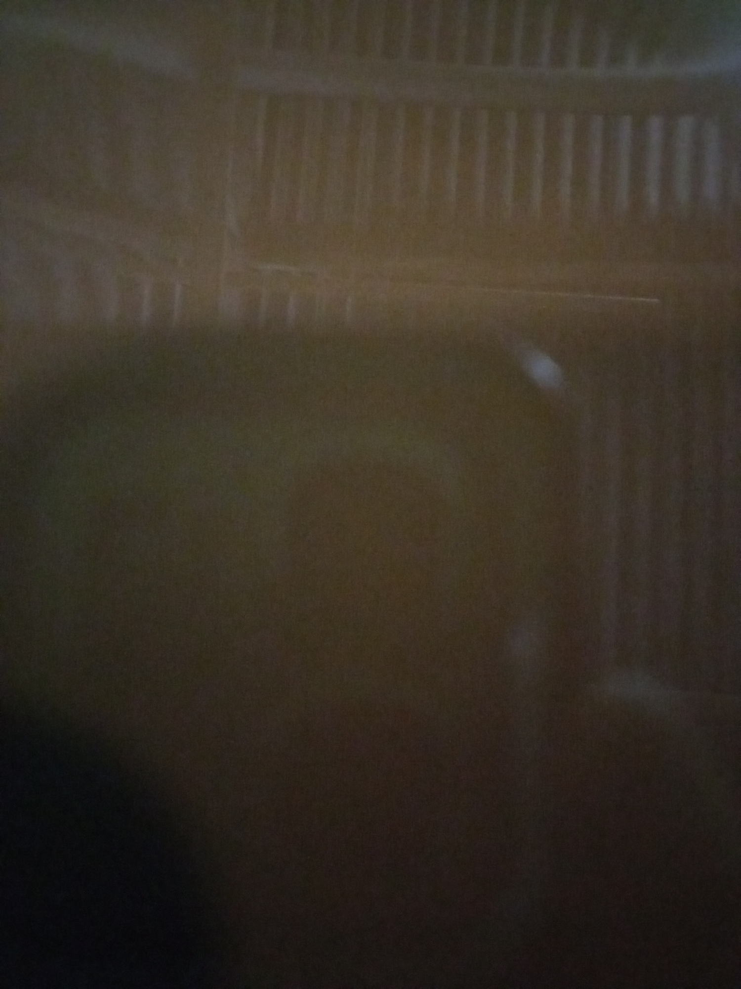 The image is a dimly lit, out-of-focus shot that appears to capture a section of a fence or grate. The bars of the fence are vertical and evenly spaced. There is a faint horizontal line across the middle of the image, possibly a railing or another structural element. The lighting is low, creating a dark and somewhat mysterious atmosphere. There is a slight blur or smudge in the upper right portion of the image, which could be a reflection or an imperfection on a lens. The overall tone of the image is muted and lacks distinct details due to the poor focus and low light conditions. No people, specific objects, or discernible activities are visible. The location context of Lhokseumawe, Indonesia, does not provide any specific visual cues within this abstract and blurry image. There is no visible text.