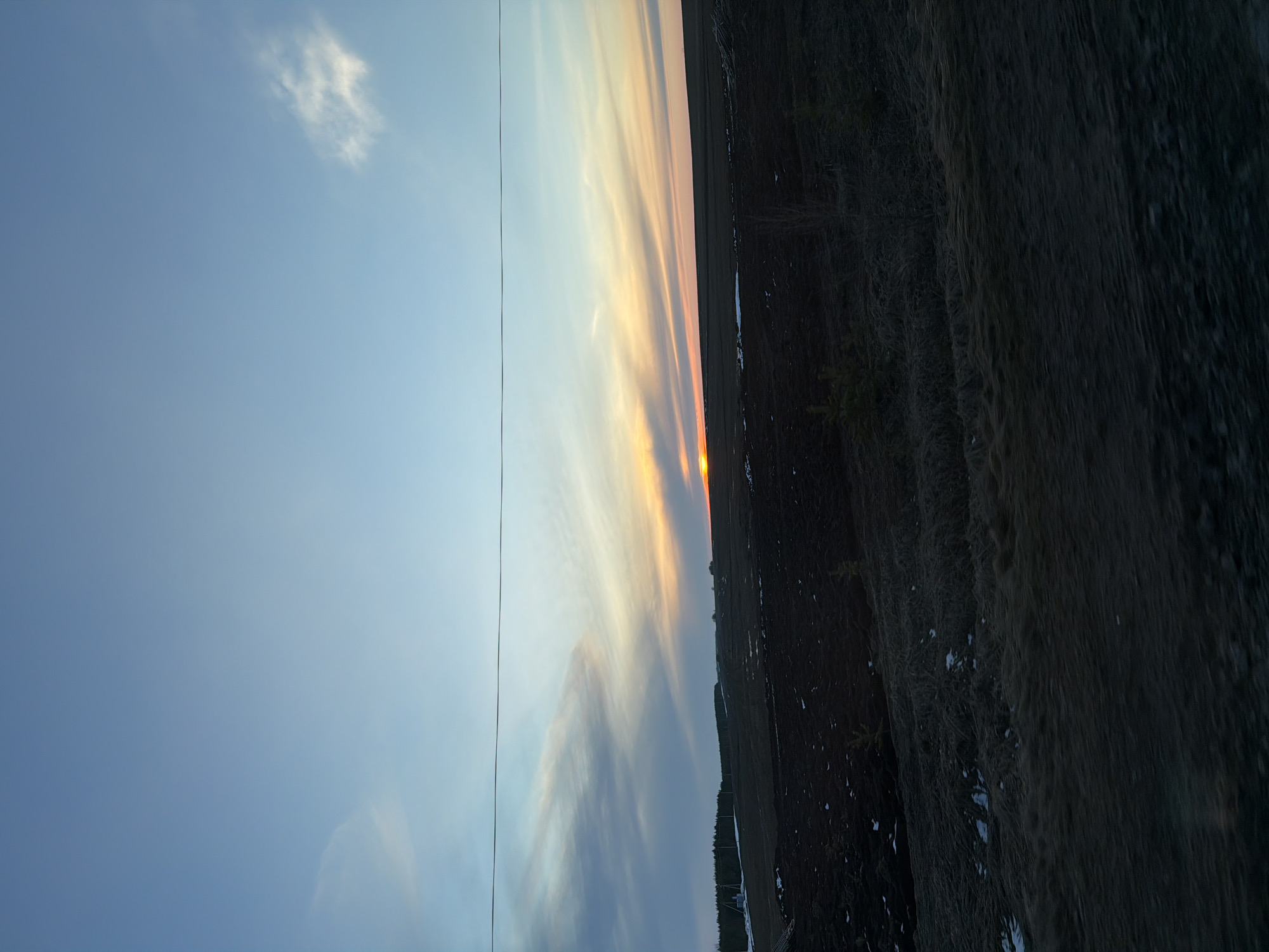The image shows a landscape in what appears to be near Дулесово, Russia based on geographic features. The sky occupies a significant portion of the image, displaying a gradient from clear blue to a sunset with orange and yellow hues. Clouds are visible in the sunset portion of the sky. A thin wire or cable stretches vertically across the left side of the image. The lower portion of the image depicts a dark, possibly barren field or land. There are some details in the field such as what seems like the beginning of trees. The scene is likely photographed at dusk or dawn.