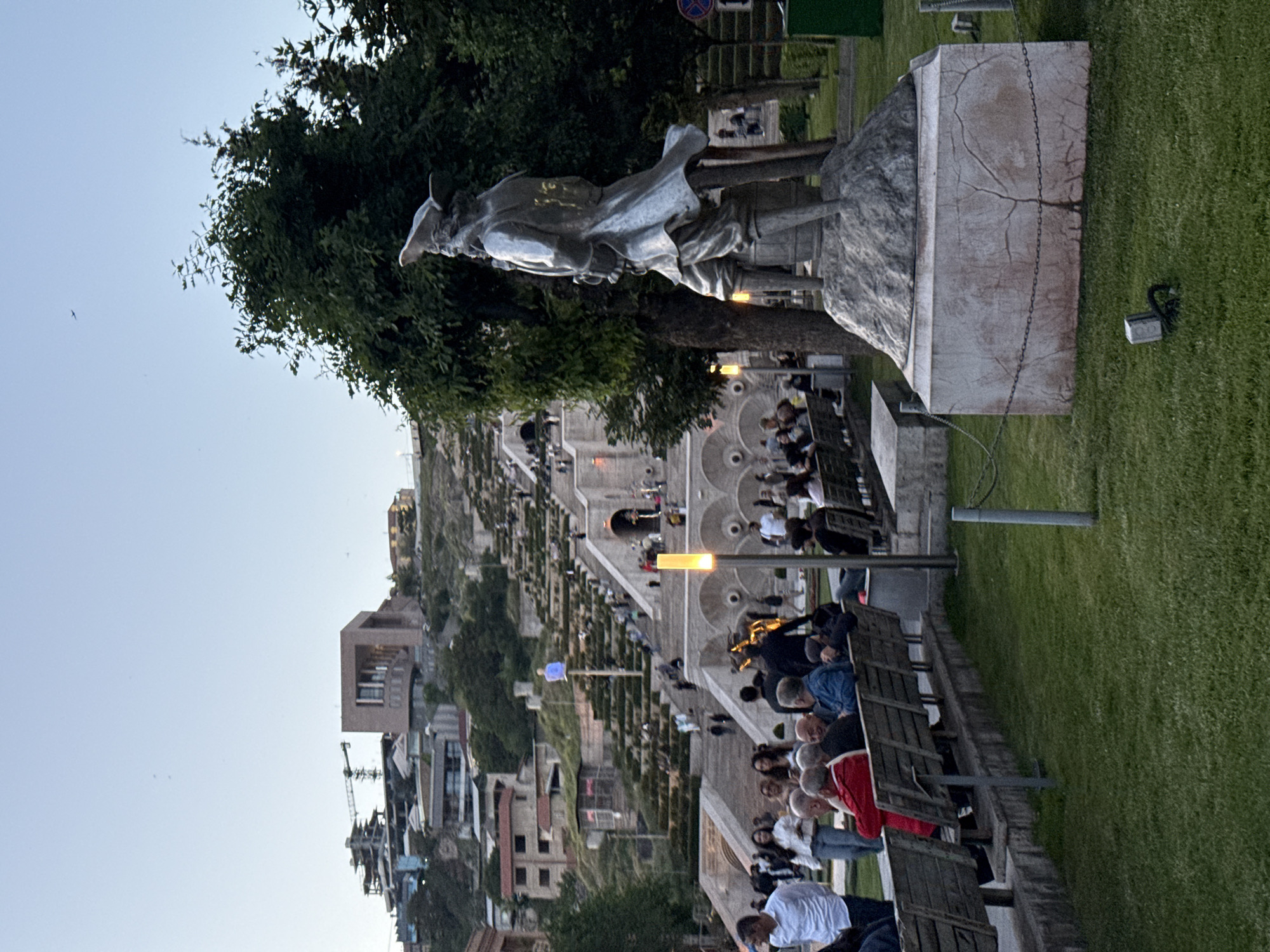 Here's a description of the image:
The image shows the Yerevan Cascade in Armenia. The composition is dominated by a large, tiered staircase with landscaping and fountains, leading up a hill. In the foreground, there are rows of wooden benches where people are sitting and observing the scene. A statue of a figure in period clothing stands on a stone plinth to the right. Lush green grass flanks the seating area. In the distance, buildings and construction cranes are visible against a pale sky.