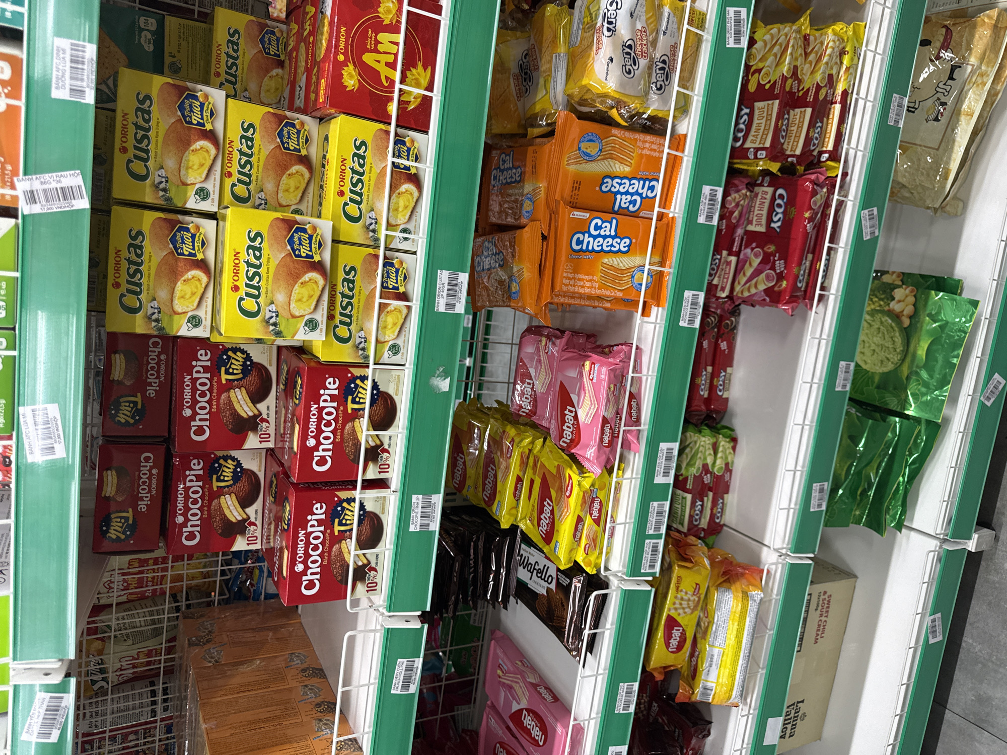 The image depicts a section of shelving within a retail environment, likely a grocery store or supermarket in Đà Nẵng, Vietnam. The shelves are stocked with various snack foods, primarily confectionery items. Brands such as Orion (Custas, ChocoPie), Cal Cheese, Nabati, Cosy and Gery are visible. Packages of wafer-style treats and cream-filled biscuits are prominent. The shelves are lined with green shelf edge labels, some displaying pricing in Vietnamese Dong (VND). The image is taken from a high angle, showing multiple shelves with a range of products. The environment appears to be well-lit and organized.