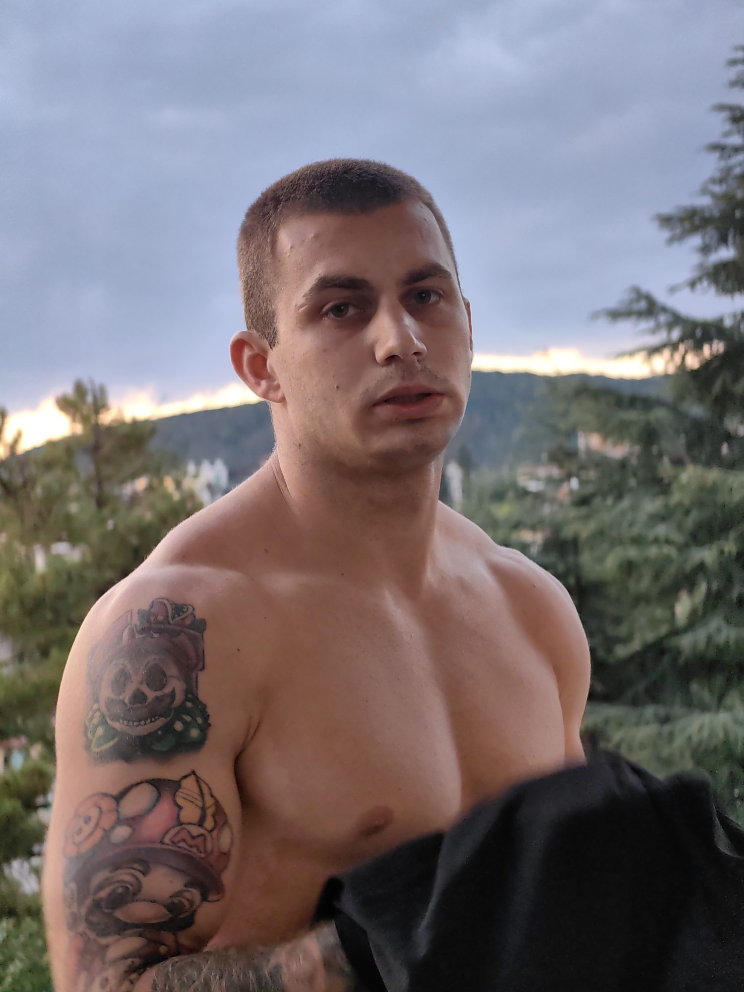 A medium shot, outdoor, vertical, eye-level photograph shows a shirtless man with short, dark hair and a fair complexion, looking directly at the camera. He has a muscular build and visible tattoos on his left arm. The man is partially obscured by a black garment held in his right hand. He stands on a balcony or elevated platform, with a blurred background of evergreen trees and hills, suggesting a location with natural surroundings within the city of Varna, Bulgaria. The sky above is overcast with hues of pale orange and blue, indicating either sunrise or sunset. The lighting is soft, creating a natural and slightly muted ambiance.