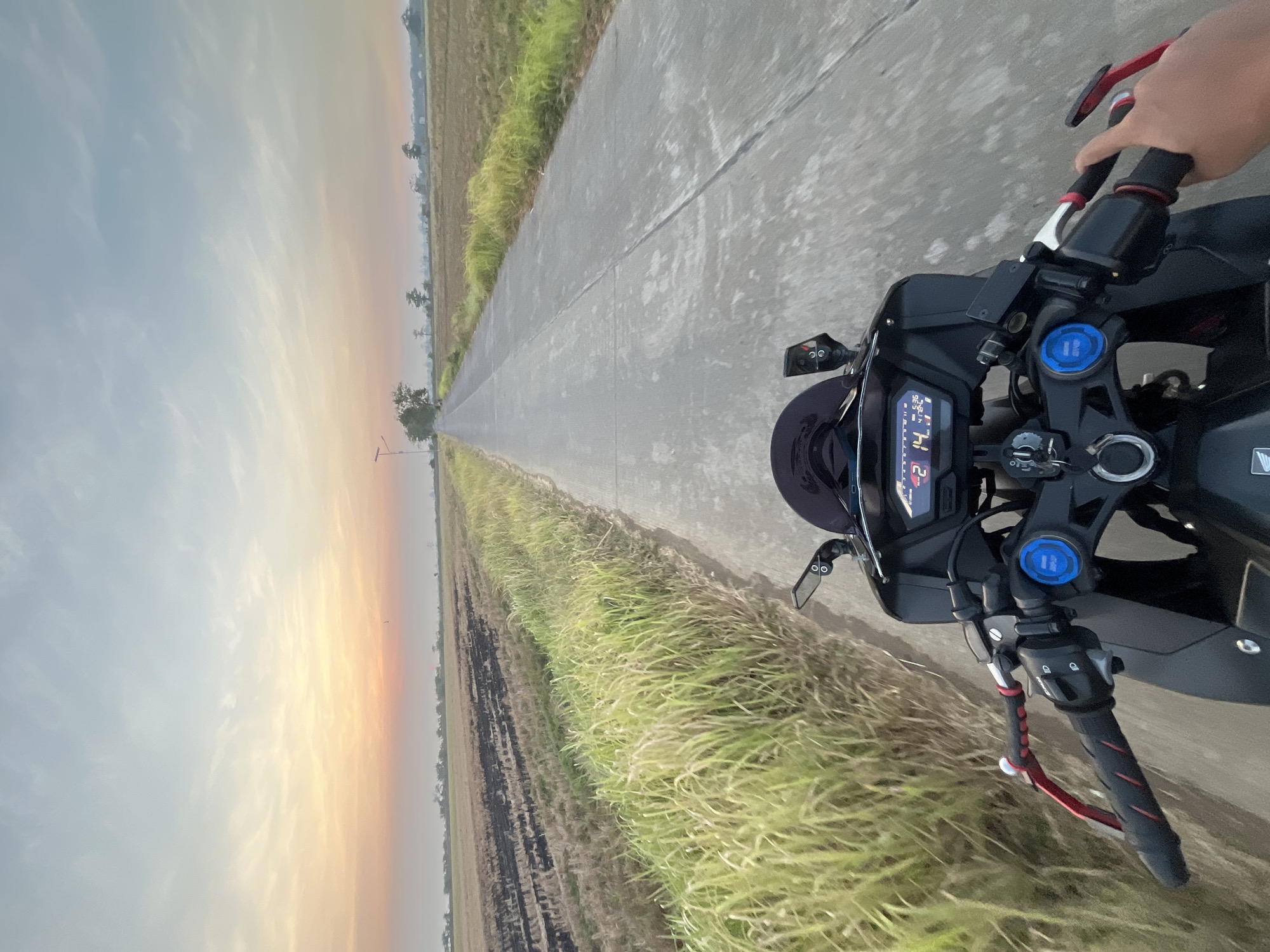 A first-person perspective captures a motorcycle ride on a paved road bordered by fields during a sunset. The sky exhibits a gradient of colors, transitioning from warm orange and pink near the horizon to a pale blue overhead, with wispy clouds. The road stretches ahead, appearing as a gray concrete path. To the left of the road, a field of dry grass or straw, some of which is scorched black, is visible, followed by taller green grass. To the right of the road, more dry field stretches out. In the distance, trees and some structures can be seen. The motorcycle's handlebars, dashboard display, and a rider's hand are prominently in the foreground. The dashboard shows various readouts, including "21 14.9 5:36 41.783". The location is specified as Pecangaan, Indonesia.