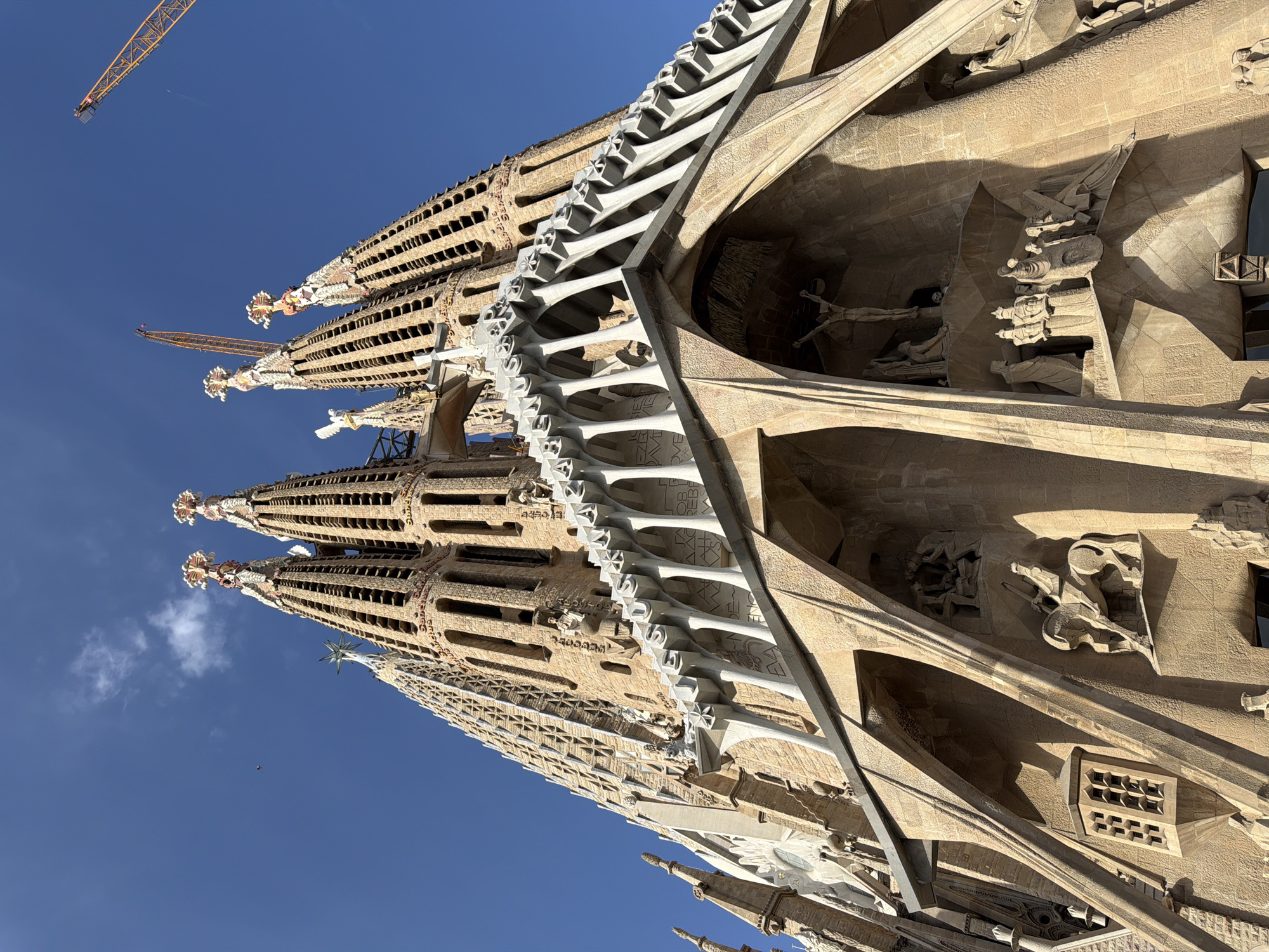 The photograph captures a low-angle view of the Sagrada Familia, a Roman Catholic minor basilica designed by Catalan architect Antoni Gaudí, located in Barcelona, Spain. The building's towering spires, adorned with intricate carvings and decorative elements, dominate the frame against a clear blue sky. The facade features a complex arrangement of arches, columns, and sculpted figures depicting religious scenes, notably a crucifixion scene prominently displayed. The natural light casts shadows, highlighting the architectural details and textures of the stone. Two construction cranes are visible in the upper right and upper center of the image, indicating ongoing construction or renovation work on the basilica. A small white object, possibly a bird or an airplane, is visible in the upper left quadrant of the sky.