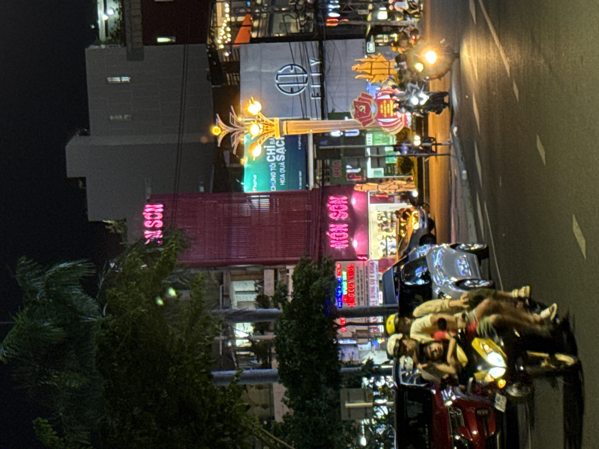 This is a nighttime photograph taken at street level, possibly in Nha Trang Ward, Vietnam. The image is oriented vertically, showing a bustling street scene. Several people are riding on motorcycles, some with passengers. The motorcycles are illuminated by their headlights and streetlights, creating streaks of light.
Buildings line the street, with neon signs and illuminated advertisements visible. One prominent sign reads "NÓN SƠN" in large pink lettering. Another sign, partly obscured by greenery, displays text that appears to be in Vietnamese, including "DỤNG CỤ Y KHOA" (medical equipment) and "HOA QUẢ SẠCH" (fresh fruit). A decorative streetlamp with multiple glowing bulbs is also visible.
The overall atmosphere is dynamic and lively, typical of a busy urban area at night. The lighting suggests artificial sources, with warm yellow and vibrant neon colors dominating the scene. The presence of multiple motorcycles and people suggests a common mode of transportation and social activity in the area. The time of day is clearly night, indicated by the dark sky and the prominence of artificial lights.