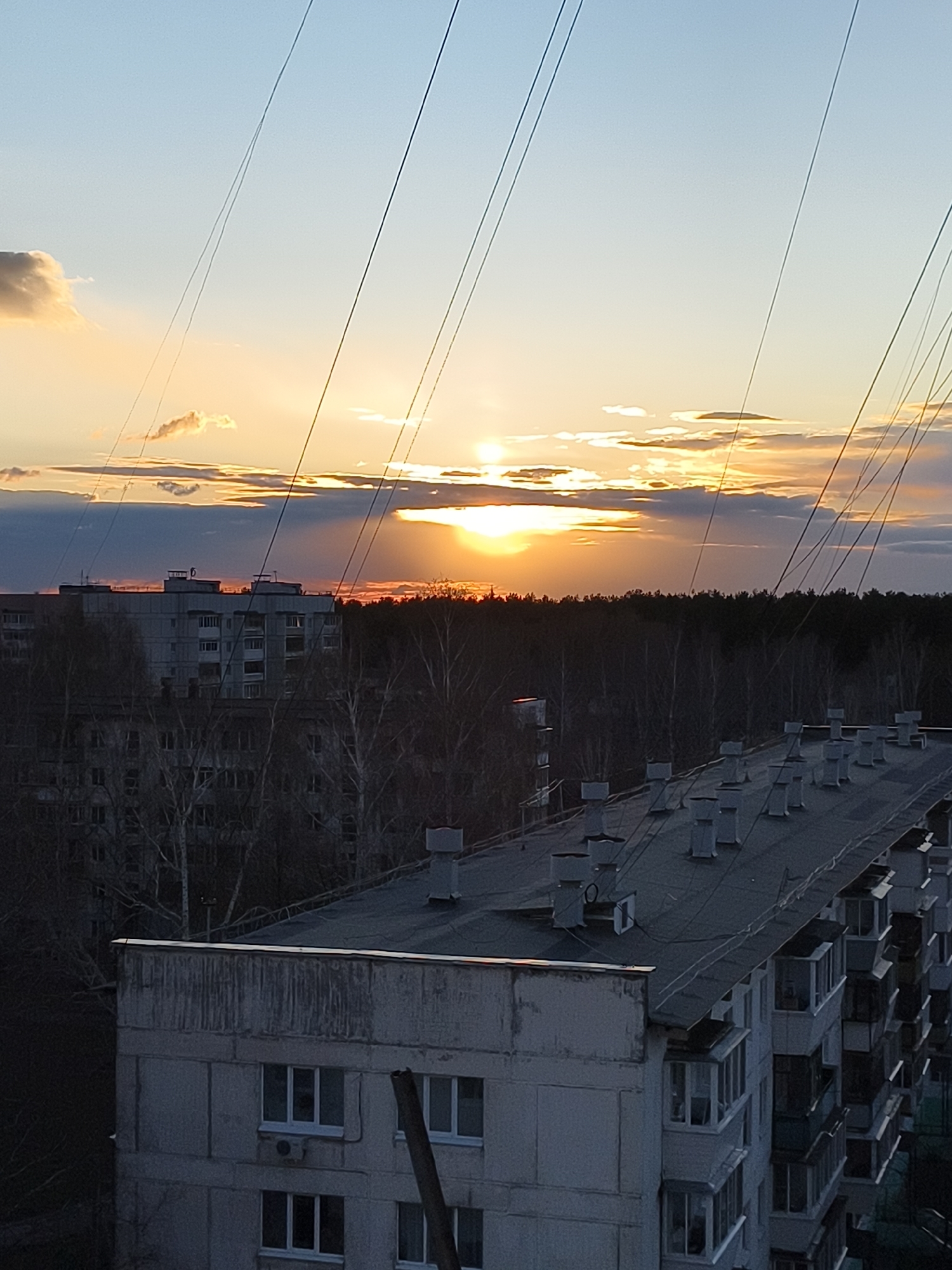 The image captures an urban landscape at sunset, viewed from a high vantage point. The scene is dominated by a dramatic sky featuring a low-lying sun, partially obscured by clouds, casting intense orange and yellow light across the horizon. Distinct sunrays pierce through breaks in the clouds.
In the foreground, the dark, flat roof of a large apartment building is visible, equipped with numerous rectangular vents or chimneys. Part of the building's facade, characterized by its grey concrete appearance and white-framed windows and balconies, extends down the right side of the frame. Several thin, dark cables or wires diagonally traverse the upper sky, originating from an unseen point close to the viewer.
The mid-ground features a dense expanse of bare, deciduous trees, forming a dark silhouette against the brighter sky. Beyond this wooded area, another large, grey apartment building or complex is discernible, partially shrouded in shadow.
No people or discernible activities are visible. The time of day is clearly sunset, as indicated by the position of the sun and the warm, fading light. The weather appears to be partially cloudy. The architecture suggests a utilitarian, possibly Eastern European or post-Soviet style. There is no visible text in the image. The overall impression is one of a serene yet somewhat stark urban environment at dusk.