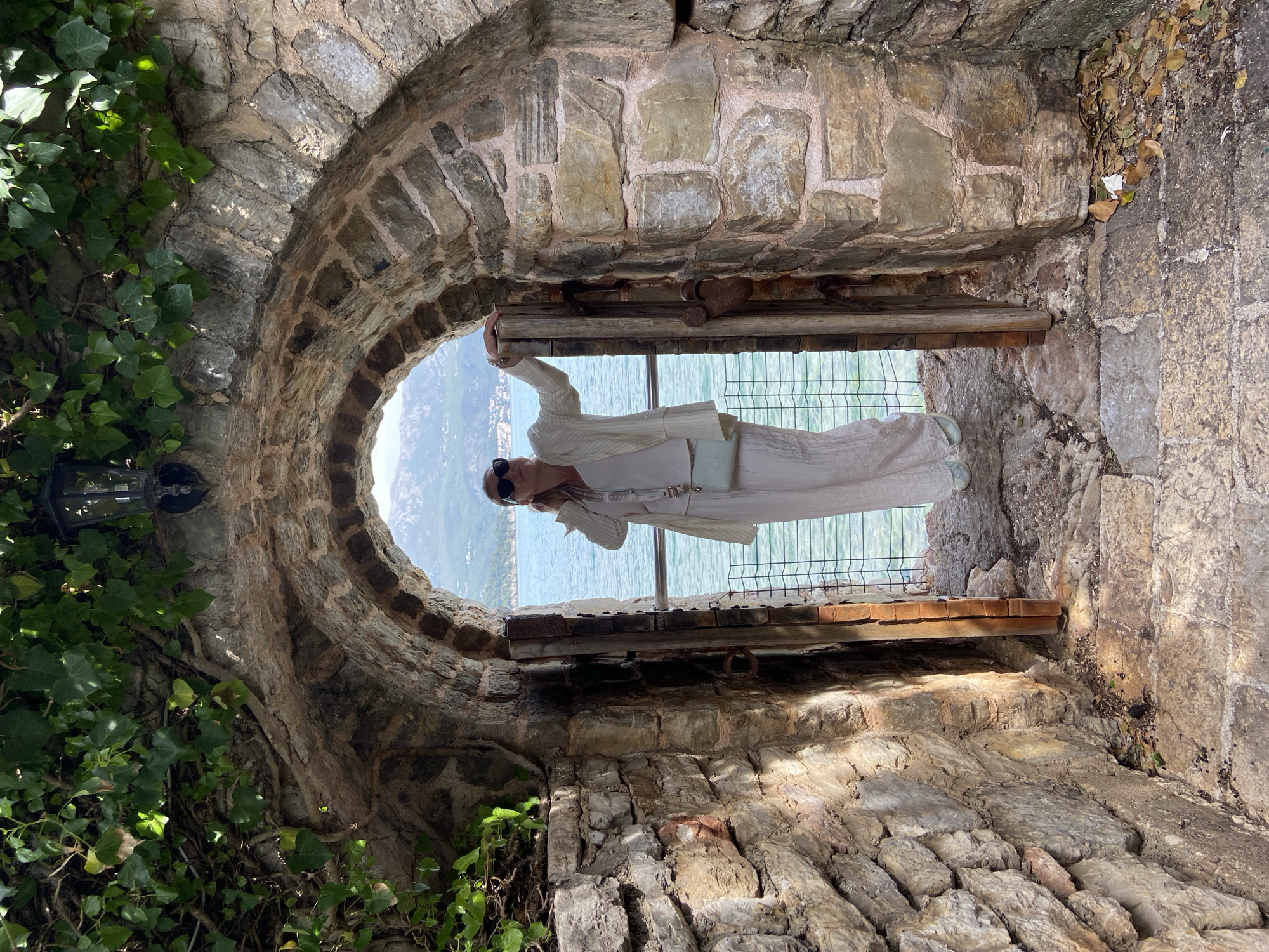 The image, rotated 90 degrees clockwise, captures a person standing within an ancient stone archway, looking out over a scenic coastal landscape. The setting appears to be a historic stone structure, fitting the provided context of Budva, Montenegro.
The central subject is a woman, dressed in an all-white or cream outfit consisting of wide-leg pants, a top, and a lightweight cardigan or jacket. She wears dark sunglasses and carries a light-colored handbag with "CK" (Calvin Klein) visible on its front. She stands framed by the archway, holding onto its stone sides, seemingly posing for a photograph or enjoying the expansive view.
The archway is constructed from large, irregularly shaped, weathered stone blocks, indicative of an old structure. To the left, lush green ivy vines with numerous leaves cling to the stone, partially obscuring a dark, possibly metallic, lantern or lamp. On the right, some dried leaves and debris are visible on an upper ledge of the stone frame. A sturdy wooden beam or shelf runs horizontally across the lower section of the opening.
Beyond the archway, a wide expanse of serene, blue-green water—likely the sea or a large lake—dominates the background. Distant landmasses or mountains rise faintly on the far horizon under a bright sky. A metal mesh fence or railing is installed across the opening, serving as a protective barrier between the person and the view. The scene is bathed in bright daylight, suggesting a sunny day, likely mid-day or afternoon. The overall impression is one of historical charm combined with natural beauty.