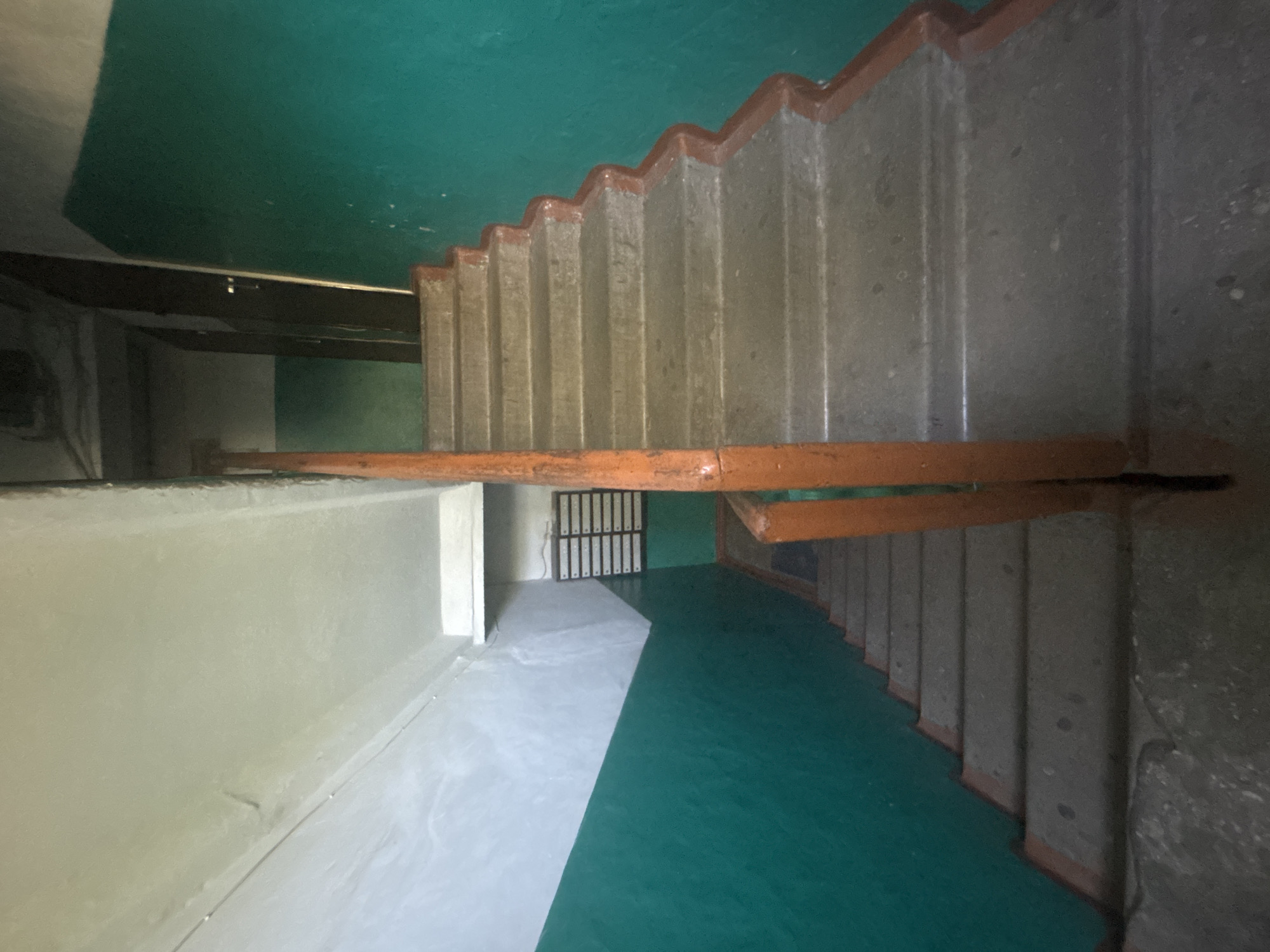 The image shows a view looking down a concrete staircase inside a building, likely an apartment building. The walls are painted with a teal green color on the lower half and white on the upper half. A wooden handrail, painted orange, runs along the right side of the stairs. On the left side, a portion of a wall with a mailbox unit is visible. The lighting suggests an indoor environment, possibly with natural light coming from an opening at the top of the stairs. The overall condition of the staircase appears worn, with some scuff marks and imperfections visible on the concrete steps. The scene depicts a common area within an older residential building.