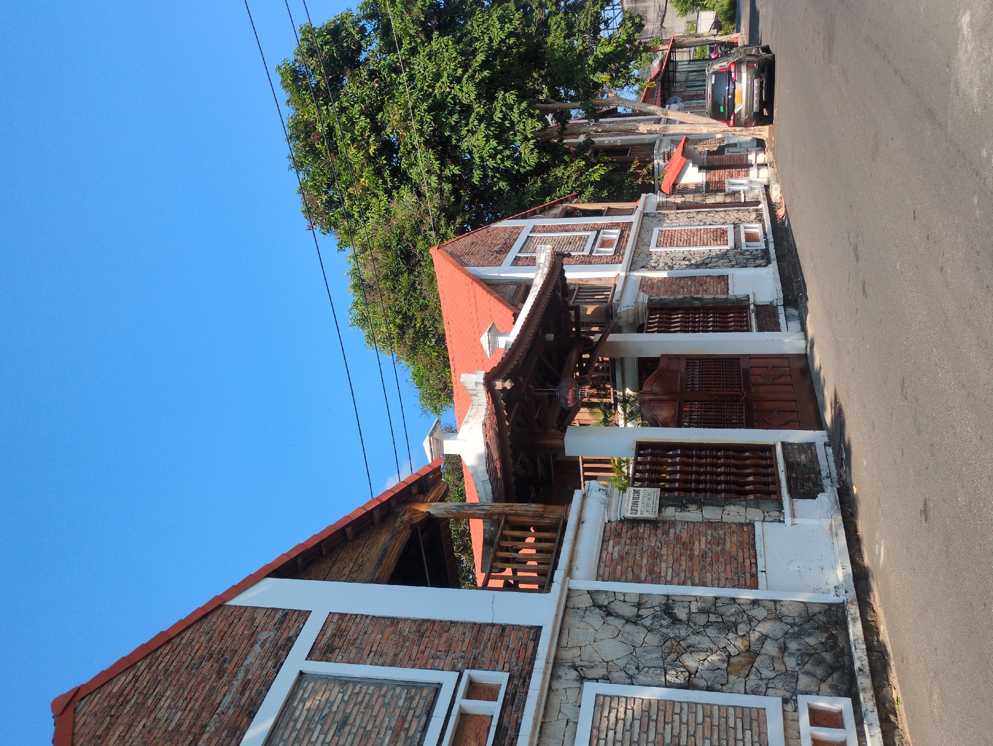 The image shows a two-story building with red-tiled roofs and brick and stone facade along a narrow road, possibly in Phú Quốc, Vietnam. The building has white trim around the windows and along the edges of the structure, with a wooden gate and fencing. Overhead power lines are visible against a clear blue sky. A large tree is to the side of the building. A car is parked on the roadside in the background. A sign is visible on the building, partially obscured, and appears to have text in English and numbers. The road appears to be asphalt. The image is taken during daylight hours with bright sunlight.