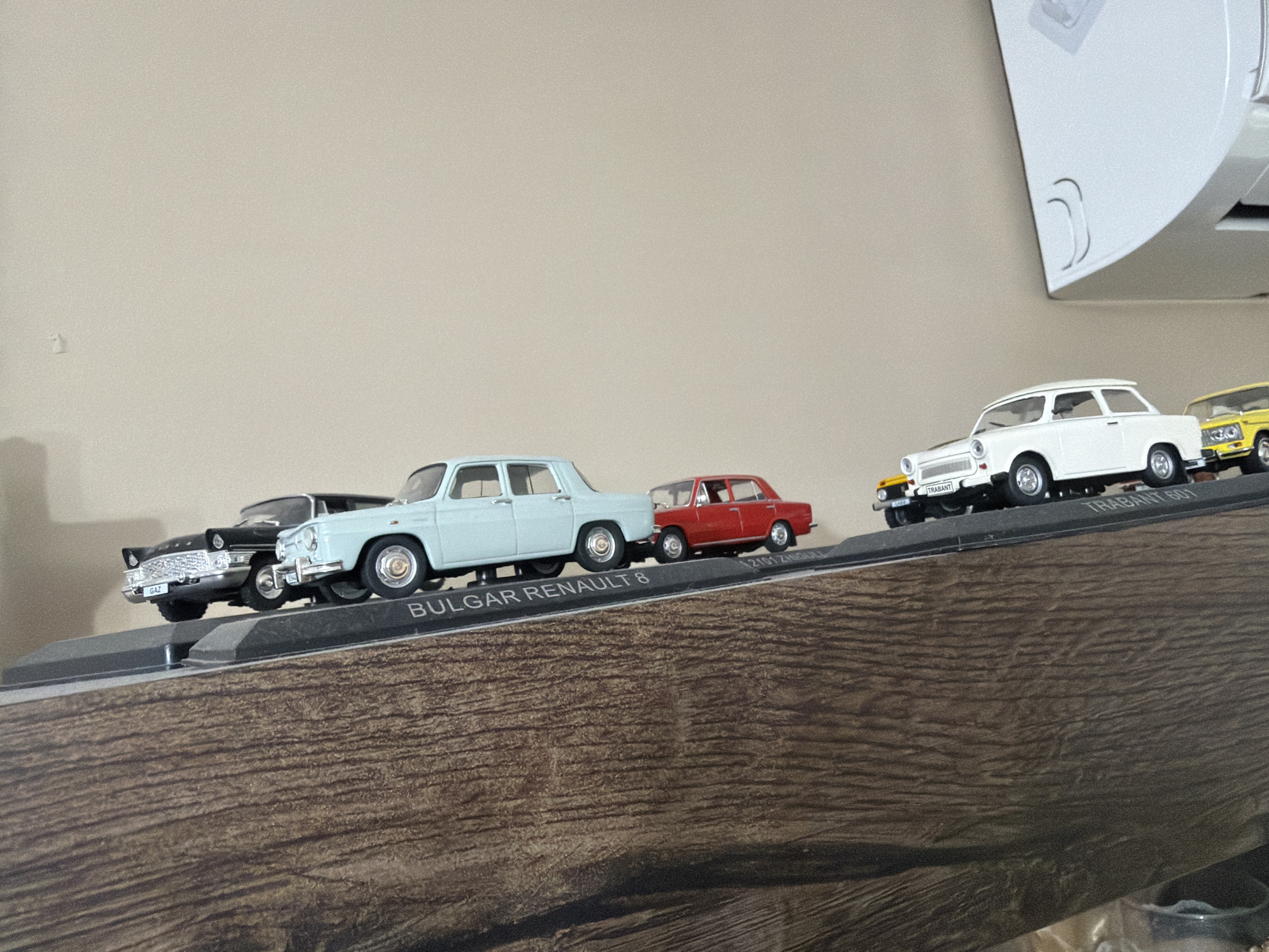 This media file shows a collection of miniature vintage cars displayed on a wooden shelf. The cars are arranged in a line, with some appearing slightly elevated relative to others, creating a dynamic display. From left to right, the visible cars include a black car with a chrome grille, a light blue sedan, a red sedan, a white hatchback, and a yellow car. Some of the cars have text on their bases, with "GAZ" visible on the black car's license plate, "BULGAR RENAULT 8" on the base of the light blue car, and "TRABANT 601" on the base of the white car. The setting appears to be indoors, with a neutral-colored wall in the background and a white, curved object, possibly part of a vent or appliance, in the upper right corner. The shelf itself has a dark wood grain texture. There are no people visible in the image, and the focus is entirely on the inanimate objects. The lighting suggests daytime, but there are no specific clues about the weather or the exact time of day. The context provided indicates the location is Burgas, Bulgaria, which might explain the "Bulgar Renault" labeling.