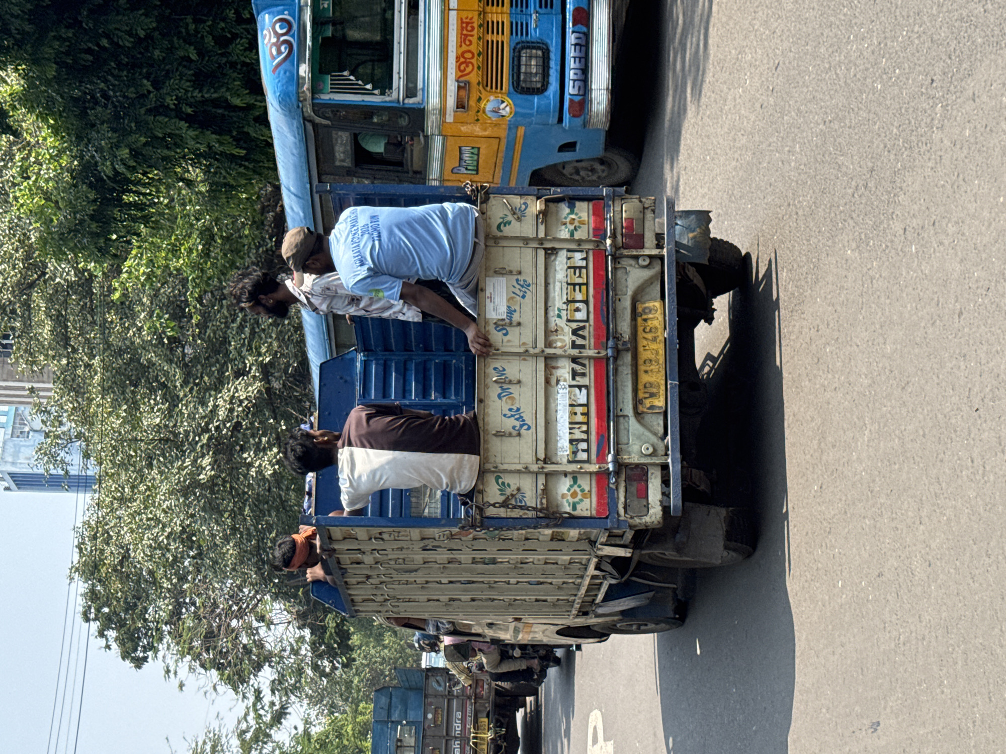 The image captures a moment on a road in Kolkata, India. A procession of blue and cream-colored trucks, possibly for transport or rally purposes, is moving along the road. At least three men are seen riding on the back of one of the trucks, with a fourth partially visible. The license plate on the truck is readable as WB 19 4619. The back of the truck also bears the text "Safe Driver" and the name "HAHETATADEEN." The text "KOLKATA POLICE MARATHON 2026 PROMO RUN" is printed on the shirt of one of the passengers. Other vehicles are following behind, indicating a moving convoy. Trees line the side of the road, suggesting a location within the city but possibly on its outskirts due to the vegetation.