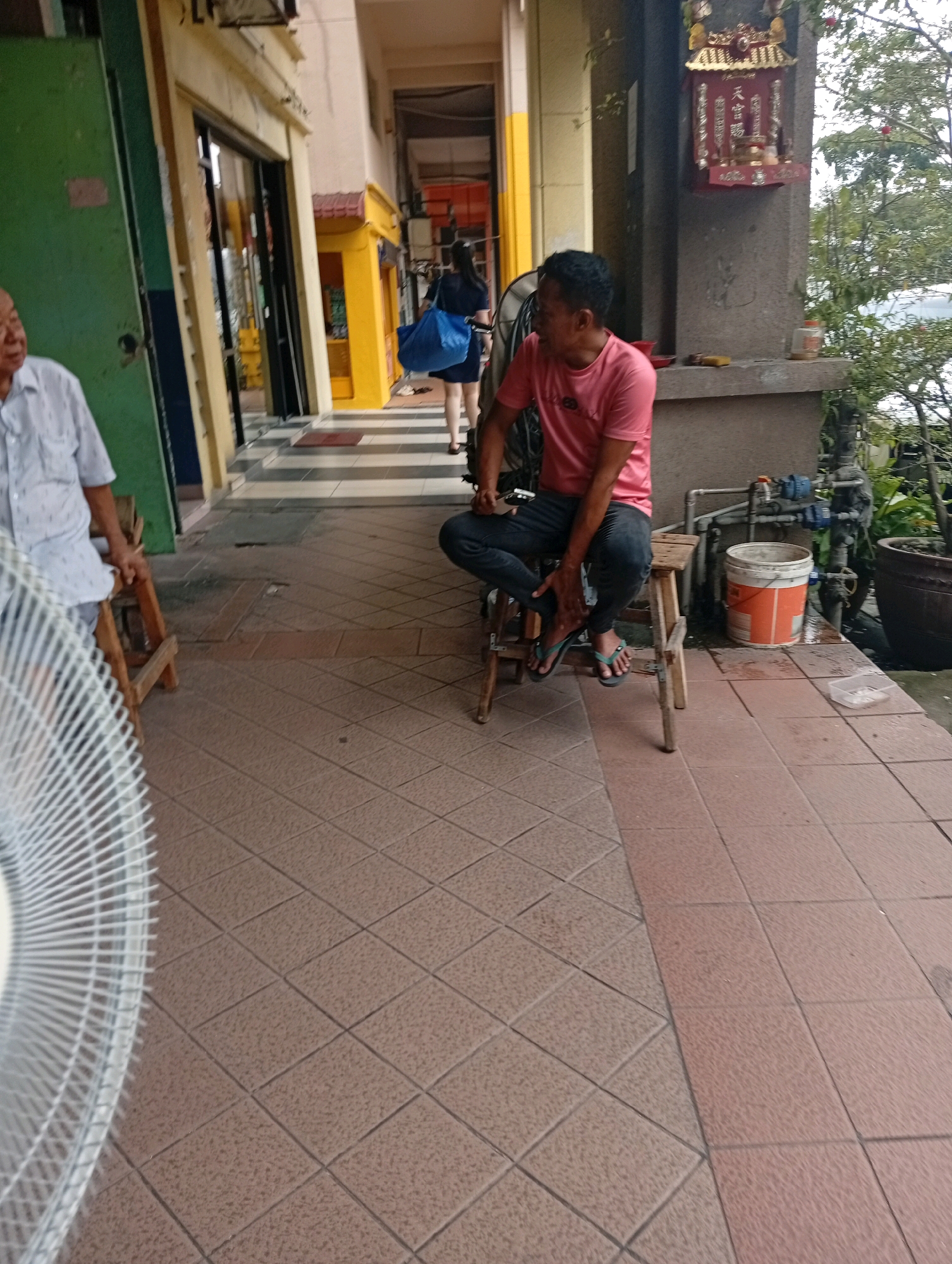 The image depicts an outdoor scene in what appears to be a covered walkway, possibly in the city of Selayang Municipal Council, Malaysia. In the foreground, a large white electric fan is partially visible on the left. To the right of the fan, an elderly man wearing a light-colored, patterned short-sleeved shirt is seated on a wooden stool. He is looking to his left. Further down the walkway, a younger man in a pink t-shirt and dark pants is sitting on another wooden stool, facing to his left, and appears to be looking down. He is wearing flip-flops. Behind him, a woman in a dark dress is walking away, carrying a large blue bag over her shoulder. The walkway has a tiled floor with a diamond pattern. In the background, there are shops with yellow and green storefronts, and a large grey concrete pillar with a small shrine attached to it. Various objects, including a white bucket with orange markings, pipes, and plants in pots, are visible along the walkway. The overall environment suggests a public area, likely a commercial or residential complex.