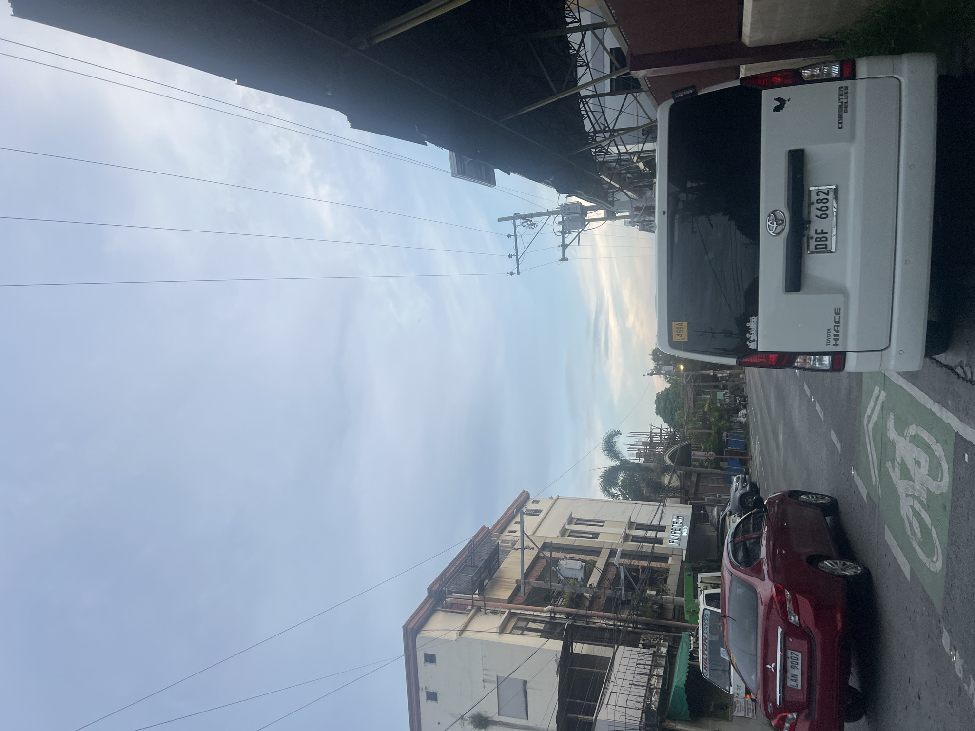 The image depicts a daytime street scene in an urban area, likely Davao City, Philippines, given the context. The perspective is from street level, looking slightly upwards, capturing a mix of vehicles, buildings, and overhead infrastructure against a light blue sky with scattered clouds, suggesting a clear day, possibly late afternoon due to the soft light.
On the right side of the frame, a white Toyota Hiace Commuter Deluxe van is prominently parked, showing its rear. Its license plate reads "DBF 6682," and a yellow sticker with "459A" is visible on the rear window. A distinctive rooster decal is affixed to its tailgate. To the left of the van, a red Mitsubishi Mirage sedan is parked on the street, displaying the license plate "LAN 9007." The road surface in front of the red car features green painted markings indicating a bicycle lane.
Overhead, numerous utility poles and a dense network of power lines crisscross the sky, connecting to various buildings and infrastructure. Below these lines, the upper parts of multi-story buildings line the street. One building has a sign reading "FLORETECH," while another partially visible sign on a vehicle (possibly a jeepney or tricycle) reads "HAMILTON." In the background, further down the street, more buildings, palm trees, and what appears to be ongoing construction with scaffolding can be seen. No explicit human activity is clearly discernible, but the presence of vehicles and commercial buildings implies a bustling urban environment.
