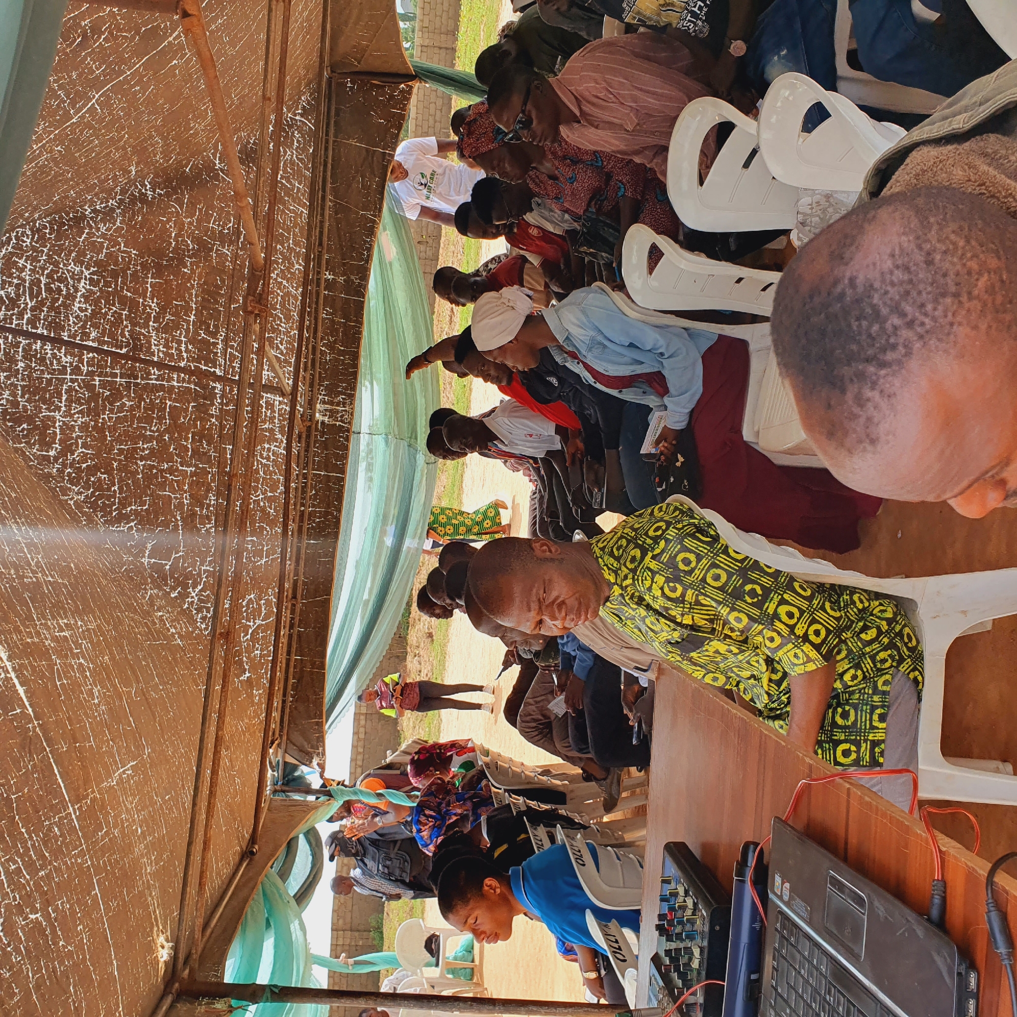 This media file depicts an outdoor gathering in Unguwar Saya, Nigeria, likely a community event or religious service taking place under a makeshift shelter. The shelter is constructed with a textured brown tarp for a roof, supported by metal poles, and draped with light green fabric on the sides.
The primary subjects are a diverse group of people, predominantly Black Africans, seated on white plastic chairs arranged in rows. Many individuals are looking downwards, suggesting they are listening or engaged in a shared activity. Some wear headscarves, indicative of local customs. The lighting suggests it is daytime, with natural light filtering through the tarp.
An individual in the foreground, facing away from the camera, wears a vibrant yellow and black patterned shirt. To their right, a laptop and audio mixer are visible on a wooden surface, implying a sound system is in use, perhaps for a speaker or musical performance.
Further back, a person in a white t-shirt with visible text stands, potentially leading or addressing the group. The text on the t-shirt appears to read "NIJAY CARE". Another person in the background wears a patterned dress. The overall atmosphere seems focused and participatory, with no overt signs of distress. The setting appears to be semi-rural, with hints of buildings and greenery visible beyond the shelter.