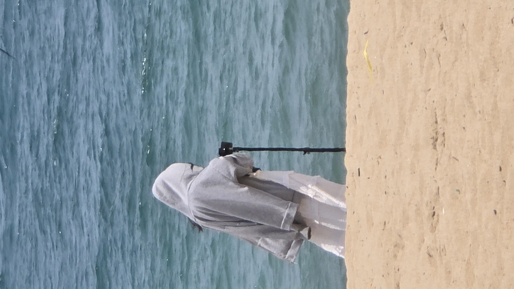 The photograph, taken at an outdoor location in Nha Trang, Vietnam, shows a person wearing a grey hooded sweatshirt and white dress standing near the edge where the sandy ground meets the blue water. A black object that could be a camera or other device is pointed towards the water. A single yellow leaf is on the ground.