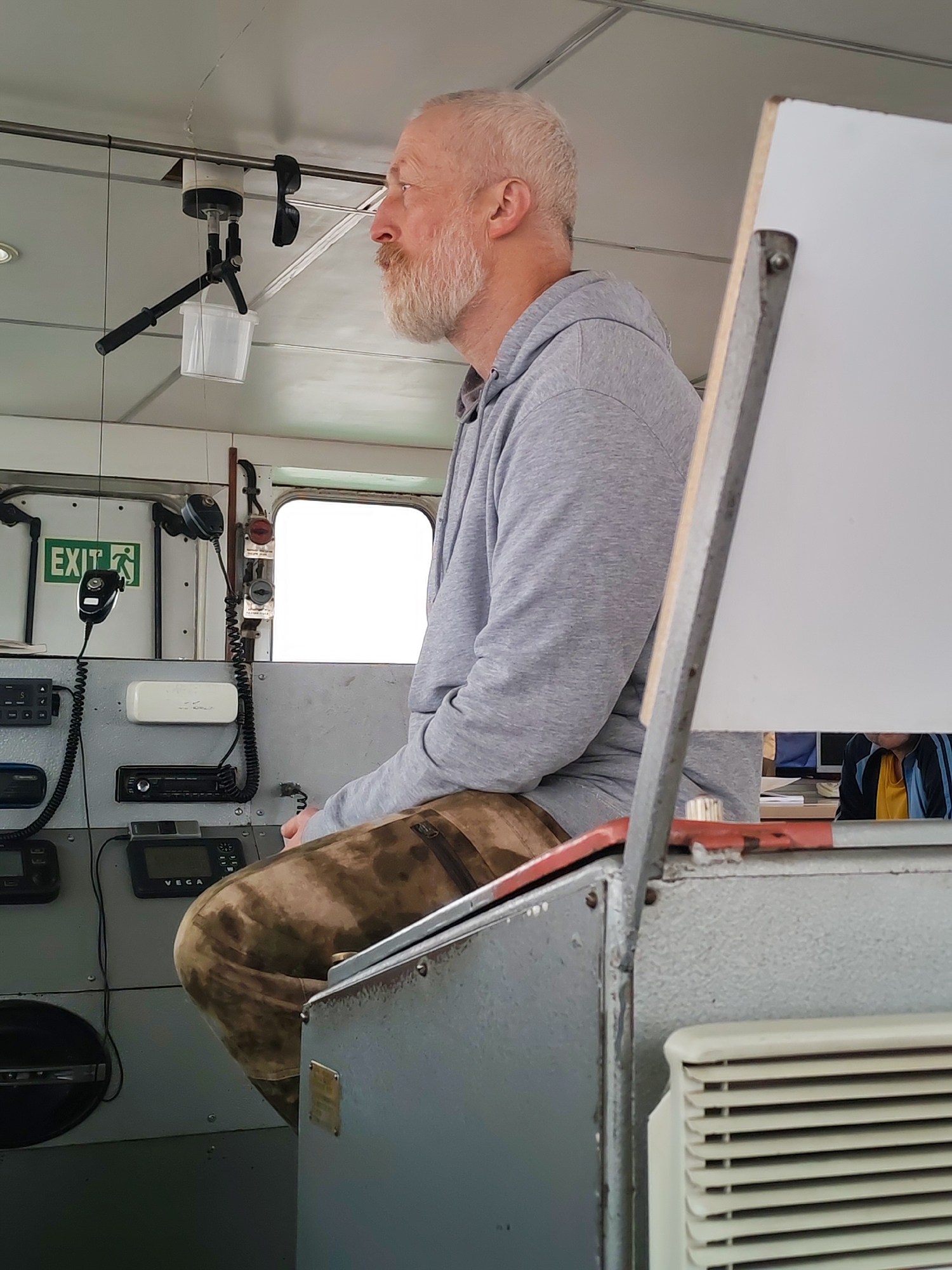 A man with a white beard and hair is seated in what appears to be the bridge or control room of a vessel, likely a ship or large boat. He is seen in profile, looking attentively towards the right side of the frame. He wears a grey hooded sweatshirt and camouflage-patterned trousers. His expression is calm and focused, suggesting he is on duty or observing something.
The setting is an interior space characterized by industrial equipment and functional design. To the man's left, a console or dashboard is visible, featuring multiple electronic devices. These include several black screens with controls, one clearly labeled "VEGA," indicating navigation or communication equipment. Two radio microphones with coiled cords hang nearby, one prominently in the foreground. A white rectangular unit and a car-style radio are also integrated into the console. Above these, an "EXIT" sign with a green background and a white running man pictogram is mounted on the wall. A bright rectangular window suggests it is daytime, showing only white light, possibly indicating an overcast sky or reflection. Two round, red-capped buttons or lights are fixed near the window.
Overhead, a metal bar runs across the ceiling. From it hangs a unique assembly featuring a small, clear plastic bucket attached to what looks like a modified tripod or mounting system. Dark sunglasses are clipped to a nearby point on the ceiling structure. To the man's right, a white panel or screen with a metal frame partially obscures the view, and a white air vent is visible at the bottom right. The overall environment suggests a working vessel, consistent with the location context of Shcheleiki, Russia, a village on a river.