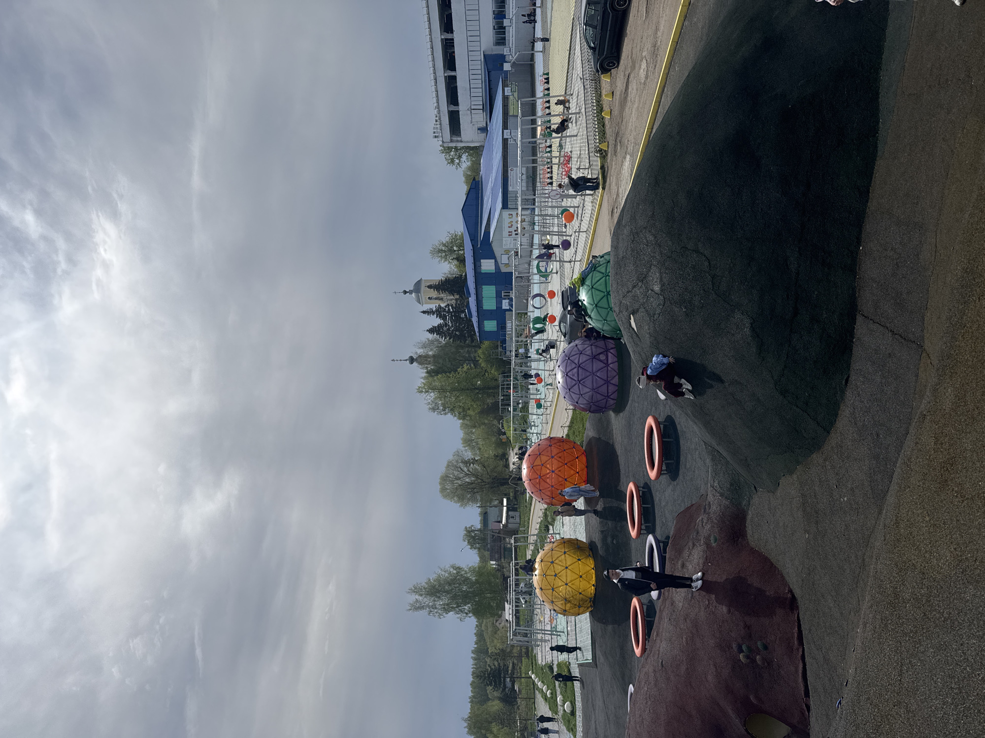 The image depicts a playground in Синьково, Russia. The playground features a sloped surface with colorful spherical climbing structures (yellow, orange, purple, and green). There are also several vertically oriented orange rings. A few people are present on the playground, including children and adults. In the background, there is a building with a blue roof and white walls. A church steeple is visible in the distance, and the sky is cloudy. A black car is parked near the building. Other people are walking near the building. The playground surface is composed of varying colors.