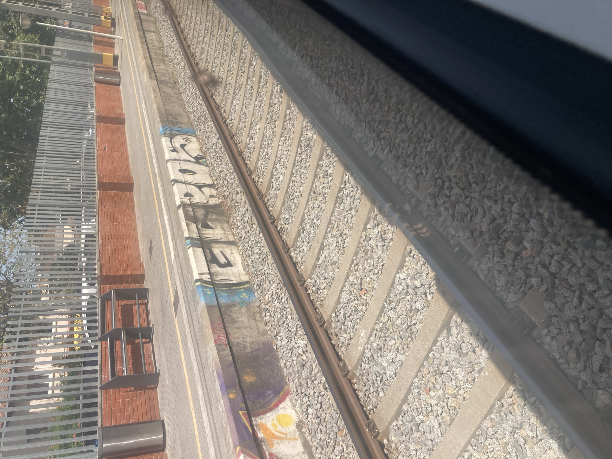 The image captures a view from a train window looking out at a train platform and tracks. The platform is lined with gravel, and a concrete barrier separates it from the tracks. Graffiti in white and blue is visible on the side of the platform barrier. Behind the platform, there is a brick wall topped with a metal fence. Two dark metal benches are situated on the platform. In the background, trees and a building are visible, suggesting an outdoor urban or suburban setting. The scene is illuminated by natural daylight. This location is identified as Cerdanyola del Vallès, Spain.