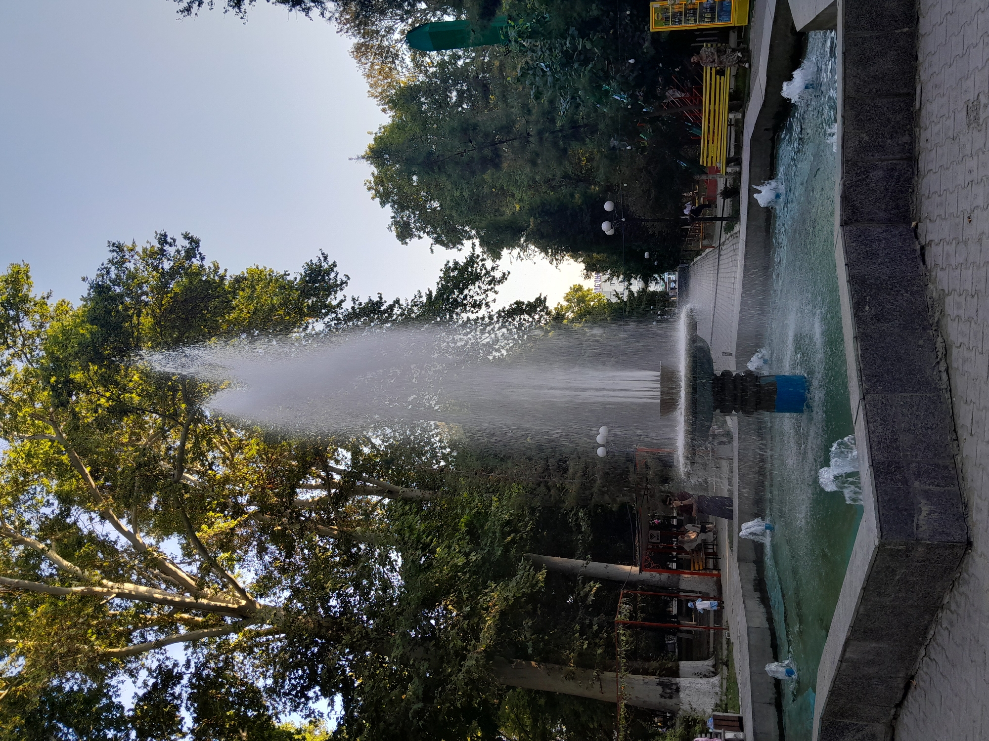 In the city of Fergana, Uzbekistan, a large, multi-tiered fountain dominates the center of the frame, with a powerful jet of water shooting high into the air. The fountain basin is hexagonal and lined with dark gray granite-like material, filled with turquoise water and smaller jets at its corners. The main part of the fountain is a dark blue and gray structure. Surrounding the fountain is a paved walkway made of interlocking gray bricks.
To the left of the fountain, large deciduous trees with light-colored bark and dense green foliage provide shade. A red and white striped canopy structure is visible, and beneath it, several people are gathered, some sitting on benches. A young child in a blue outfit is standing on the walkway.
To the right of the fountain, a pathway continues, flanked by more trees. A bright yellow bench is positioned along this path, with a person sitting on it. Further to the right, a vending machine stocked with drinks is visible, and another person is sitting on a low wall or curb nearby. The overall environment suggests a public park or plaza, likely in a warm climate given the attire and greenery. The sky is a clear, light blue, indicating daytime.