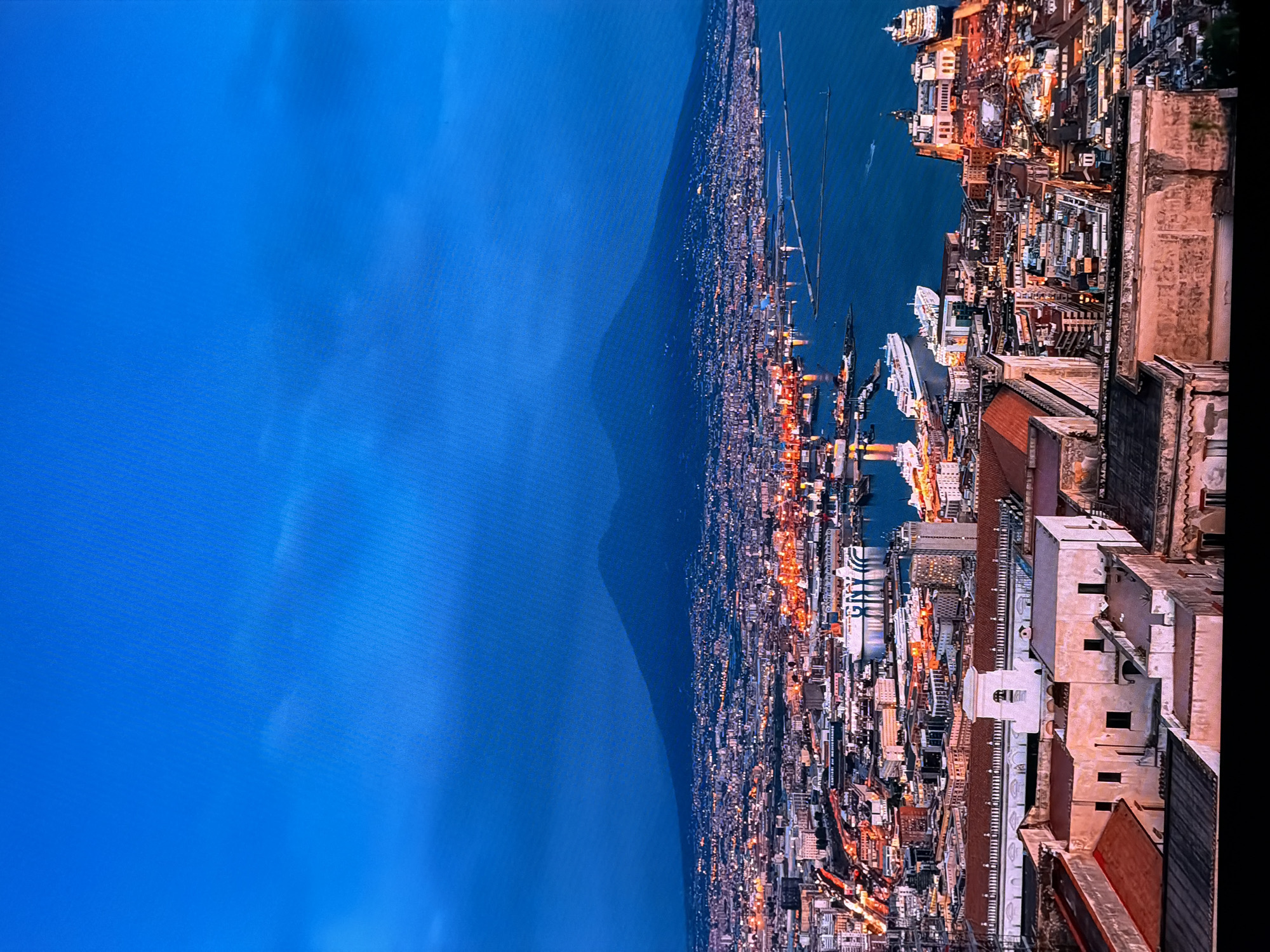 This image is an aerial view of the city of Naples, Italy, at dusk. The prominent feature in the background is Mount Vesuvius, a stratovolcano, silhouetted against the deep blue sky. The city is illuminated with countless lights, indicating it is nighttime. A large body of water, likely the Bay of Naples, is visible on the right side of the image, with several large ships docked or moving within the harbor. The foreground shows a densely populated urban area with buildings of various sizes and architectural styles, some with red-tiled roofs. The overall scene depicts a bustling cityscape at twilight, with the natural landscape of the volcano serving as a dramatic backdrop. There are no people or specific actions visible in the image.