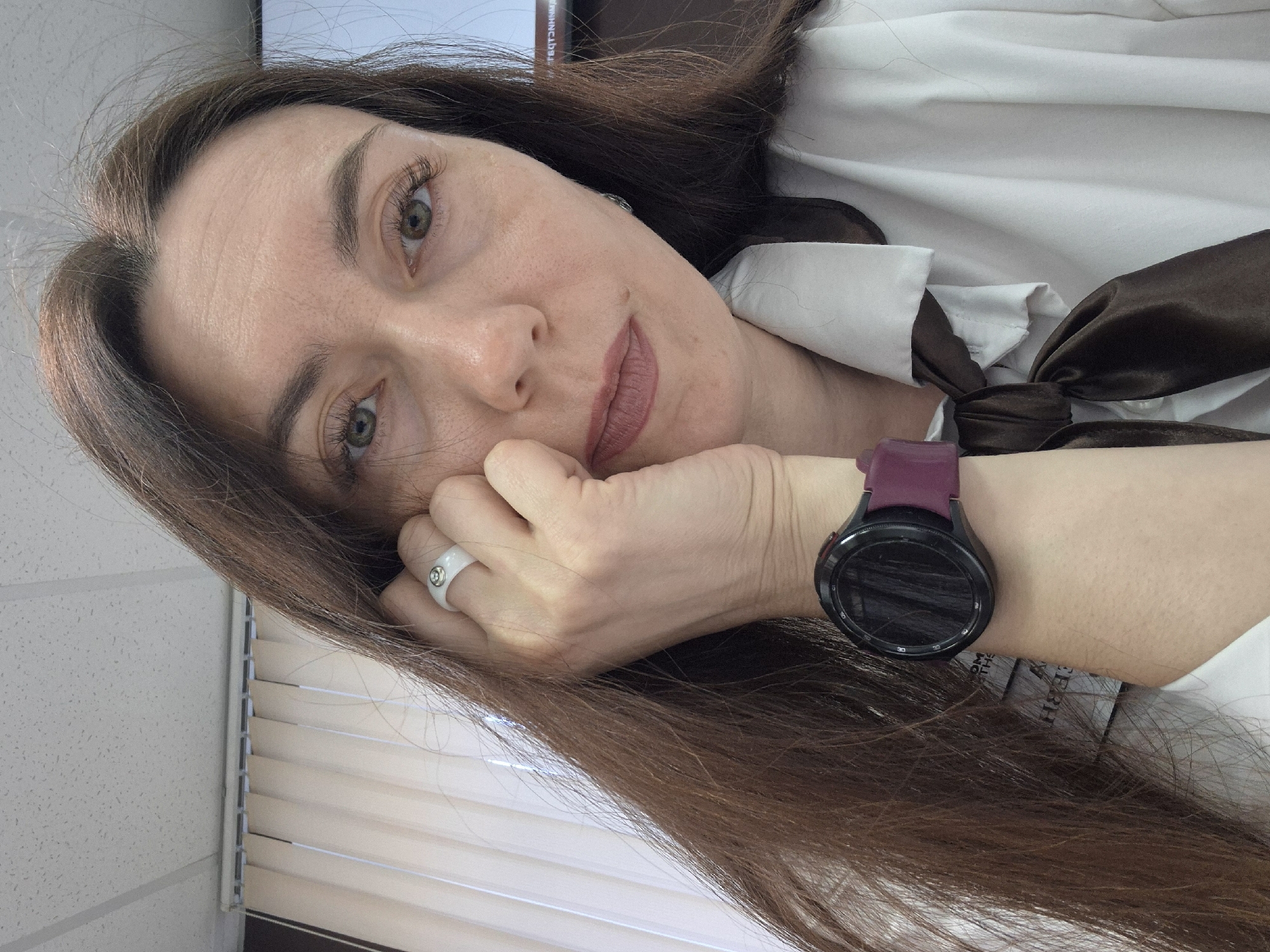 A woman is posing for a selfie indoors. She has long brown hair and is wearing a white shirt with a brown bow tie. She is wearing a black smartwatch with a purple band on her left wrist and a white ring with a small stone on her left hand. Her hand is resting on her face. Blinds are partially visible in the background. The location is in the city of Unknown, Unknown.