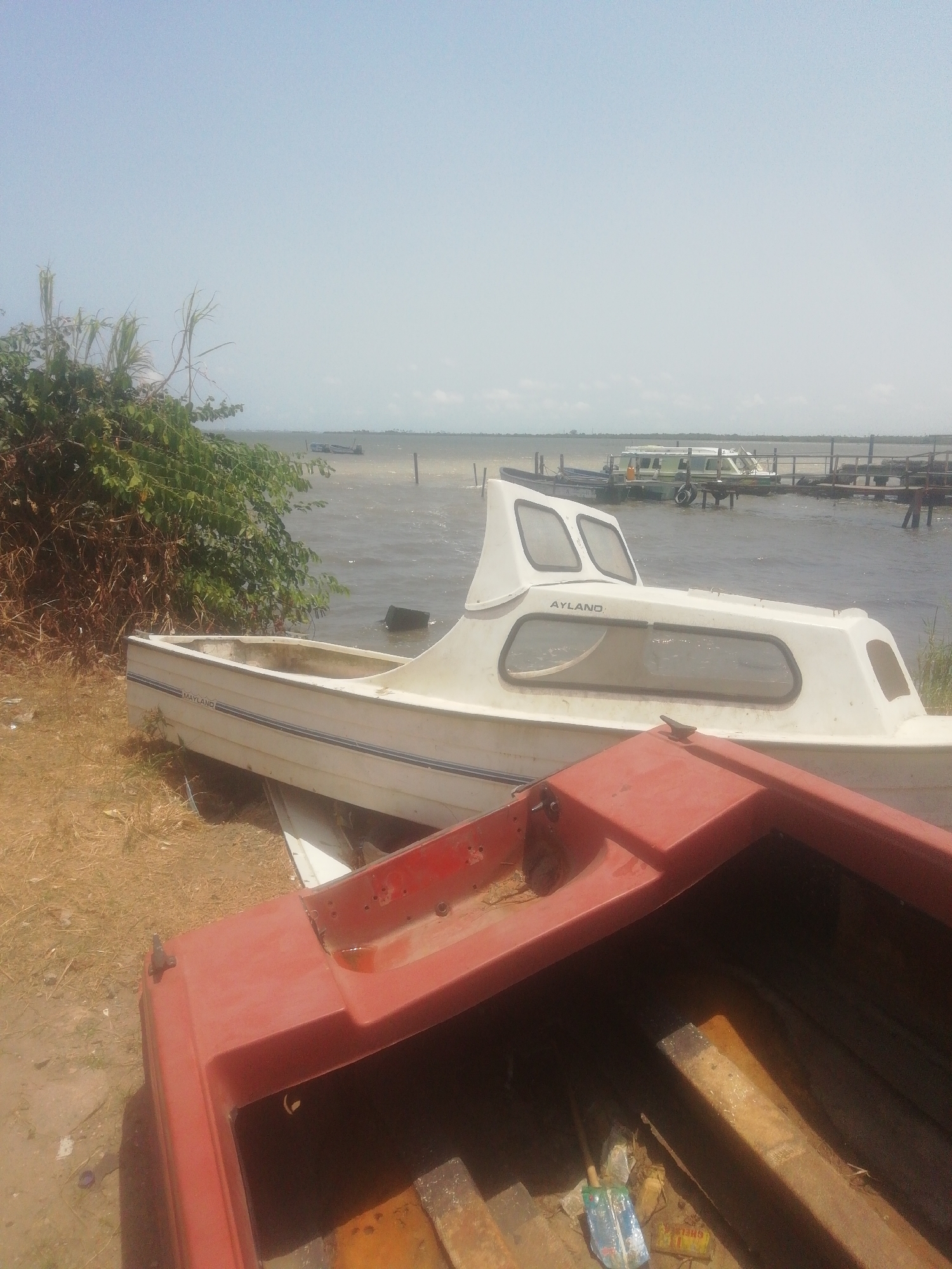 The scene shows two boats on land. The front boat is red. Behind it, a white boat named "AYLAND" is visible. The boats are located near a body of water, possibly a lagoon, with a pier extending into it. Other boats are docked at the pier. There is some vegetation nearby. Based on the prompt, the location is in the city of Ikorodu, Nigeria. No people are visible in the image.
