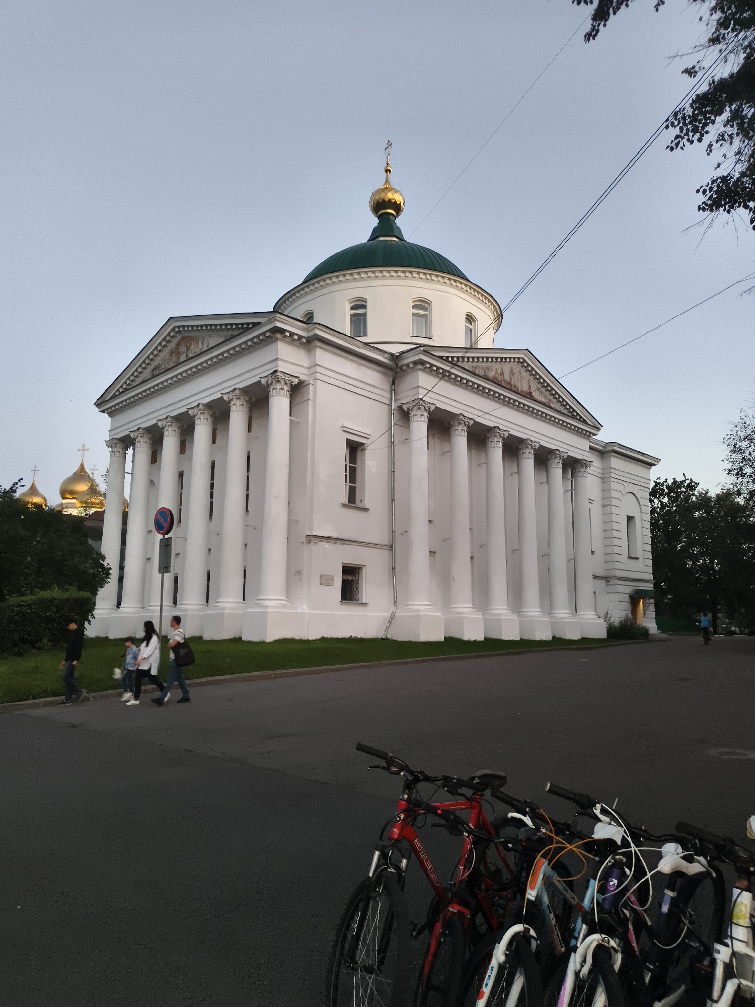 The image depicts the Church of Elijah the Prophet in Yaroslavl, Russia, a prominent architectural landmark. The white neoclassical building features a large green dome topped with a golden cross. A colonnade of white pillars supports the pediment, which has classical detailing. In the foreground, several bicycles are parked, their frames and wheels visible. A group of people are walking on the sidewalk in front of the church, with one person on a bicycle further down the street. The sky is overcast, suggesting it is either early morning or late evening. The surrounding area is a mix of greenery and asphalt.