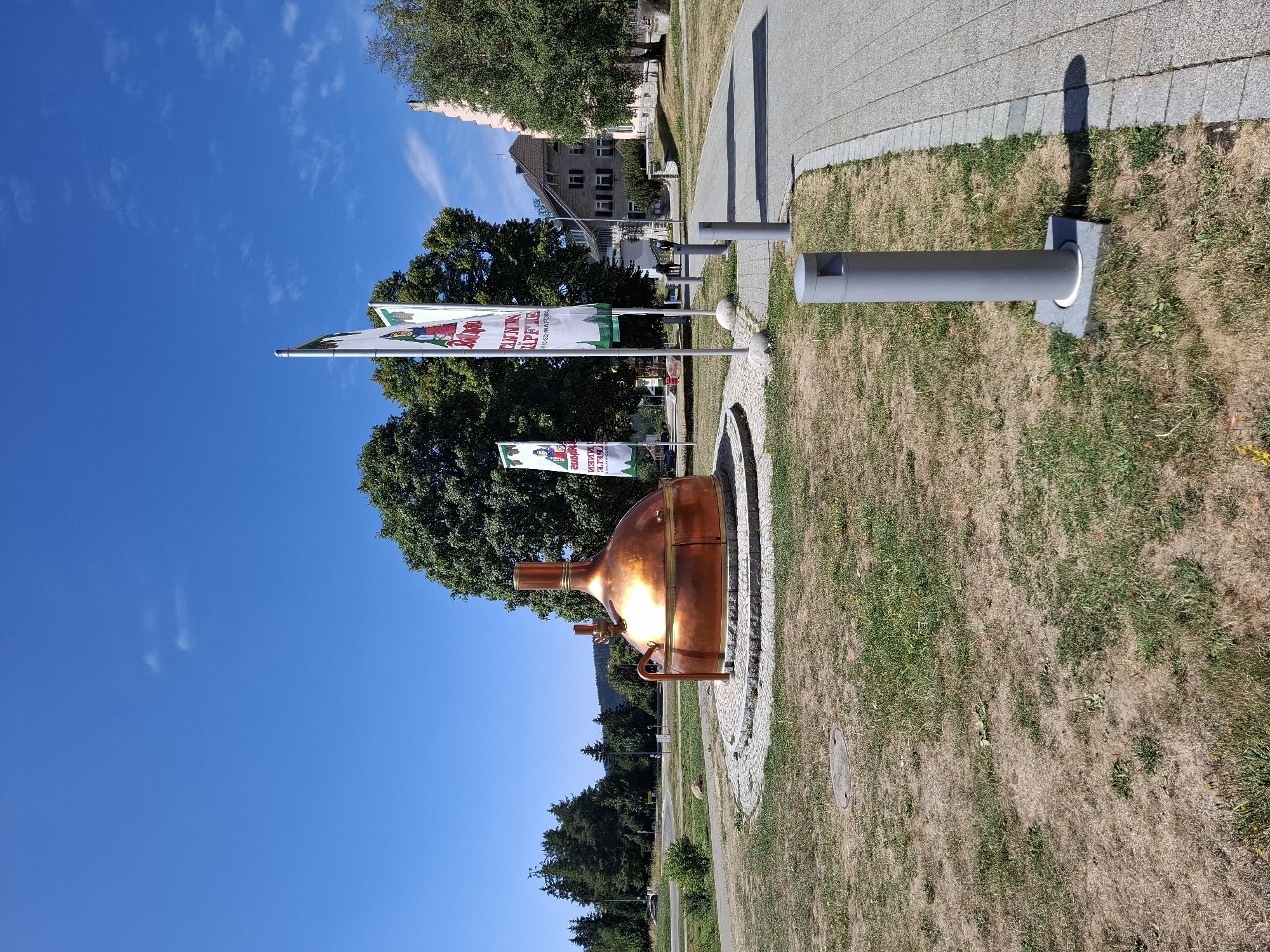 The image captures a sunlit outdoor scene in Grafenhausen, Germany. In the foreground, a large, ornate copper brewing kettle, likely a monument or decorative piece, sits on a circular stone base surrounded by dry grass. To the right, a paved walkway with grey paving stones leads into the background, bordered by a strip of grass with several modern grey bollards. Behind the kettle, three flags on poles stand tall, displaying colorful graphics and text, one of which appears to read "TANNEN ZAPFE". In the background, lush green trees dominate the landscape, with a pale pink building visible on the far right. The sky is a clear, vibrant blue with a few wispy clouds. The overall impression is one of a well-maintained public space on a bright, warm day.