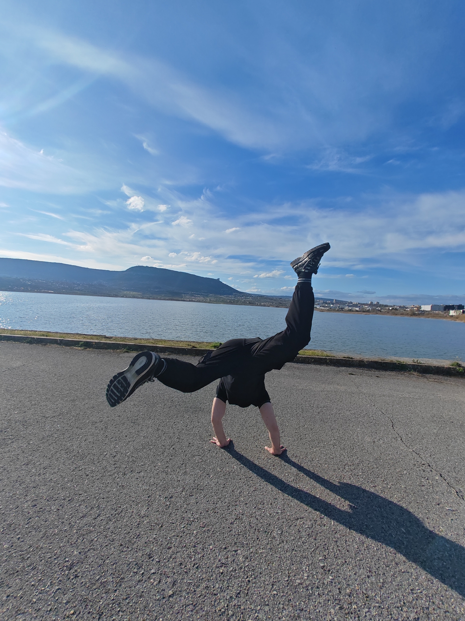 A person is performing a handstand on a paved surface, presumably a road or parking lot. They are wearing black clothing and dark-colored athletic shoes. Their shadow is visible on the pavement.
In the background, there is a body of water with a shoreline. Beyond the water, there is a mountain range and urban development which identify the location as Shumen, Bulgaria. The sky is blue with scattered white clouds.