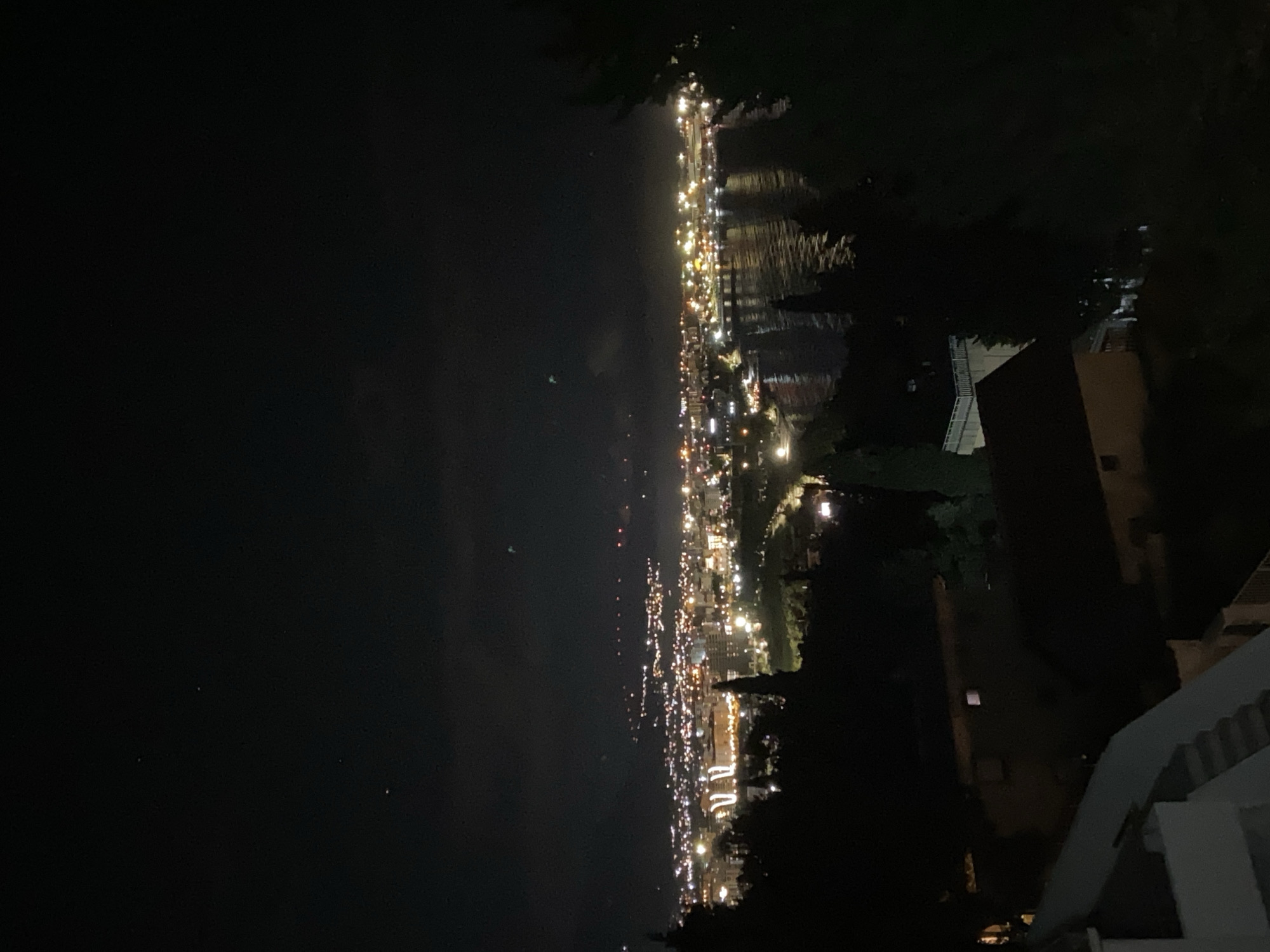 The image shows a nighttime view of the coastal city of Šušanj, Montenegro. The city lights are visible along the shoreline, reflecting in the water. The left side of the image is predominantly dark, representing the sea. On the right side, trees and buildings are partially visible, with a white building in the foreground.