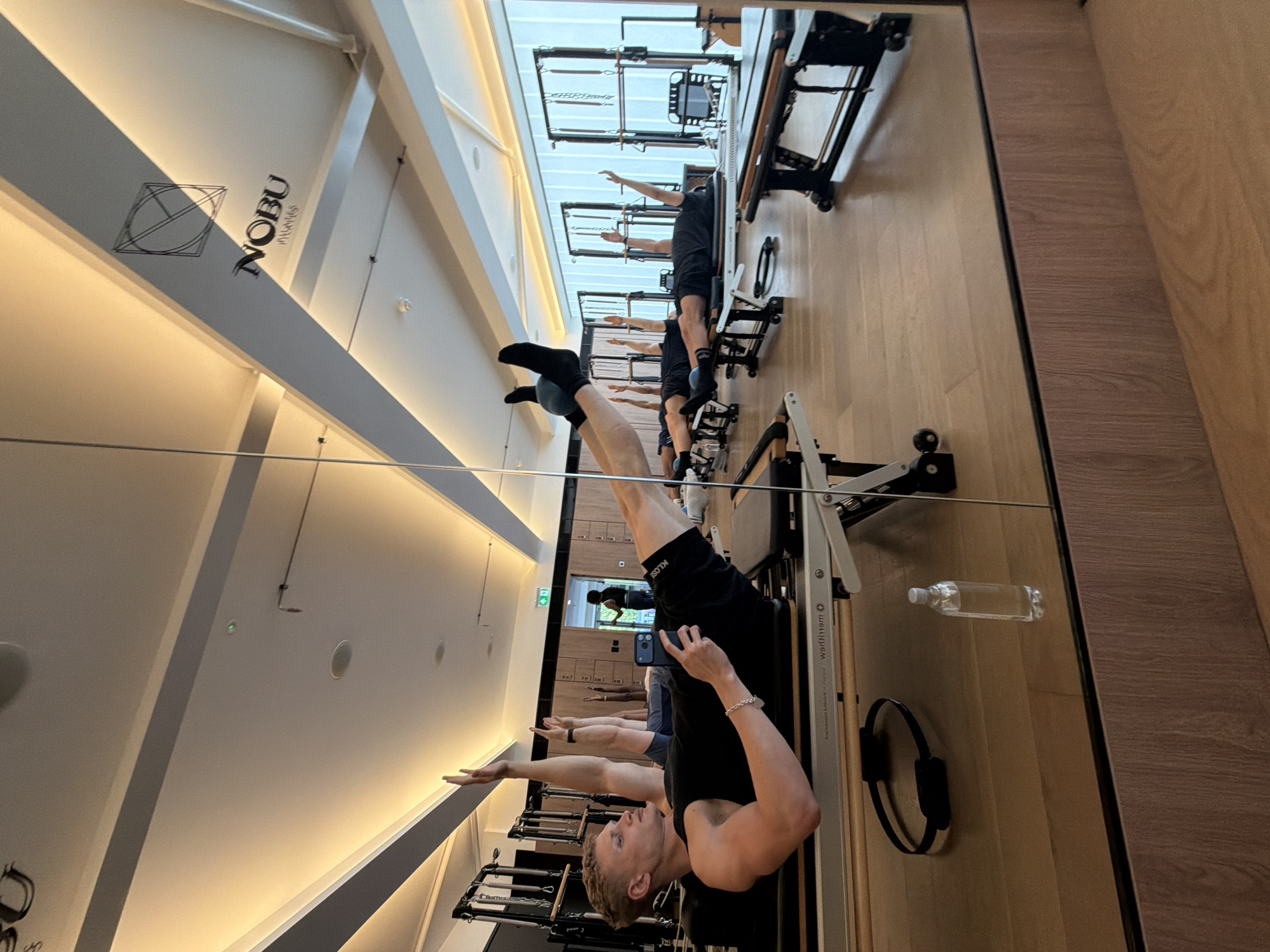 Captured in a mirror, this image showcases a group of individuals participating in a pilates session at Nobu Pilates in London, United Kingdom. The participants are utilizing pilates reformer machines in a well-lit studio environment, as indicated by the "NOBU Pilates" logo on the ceiling. The studio features a modern interior design. One participant is holding a phone, potentially taking a photo or video.
