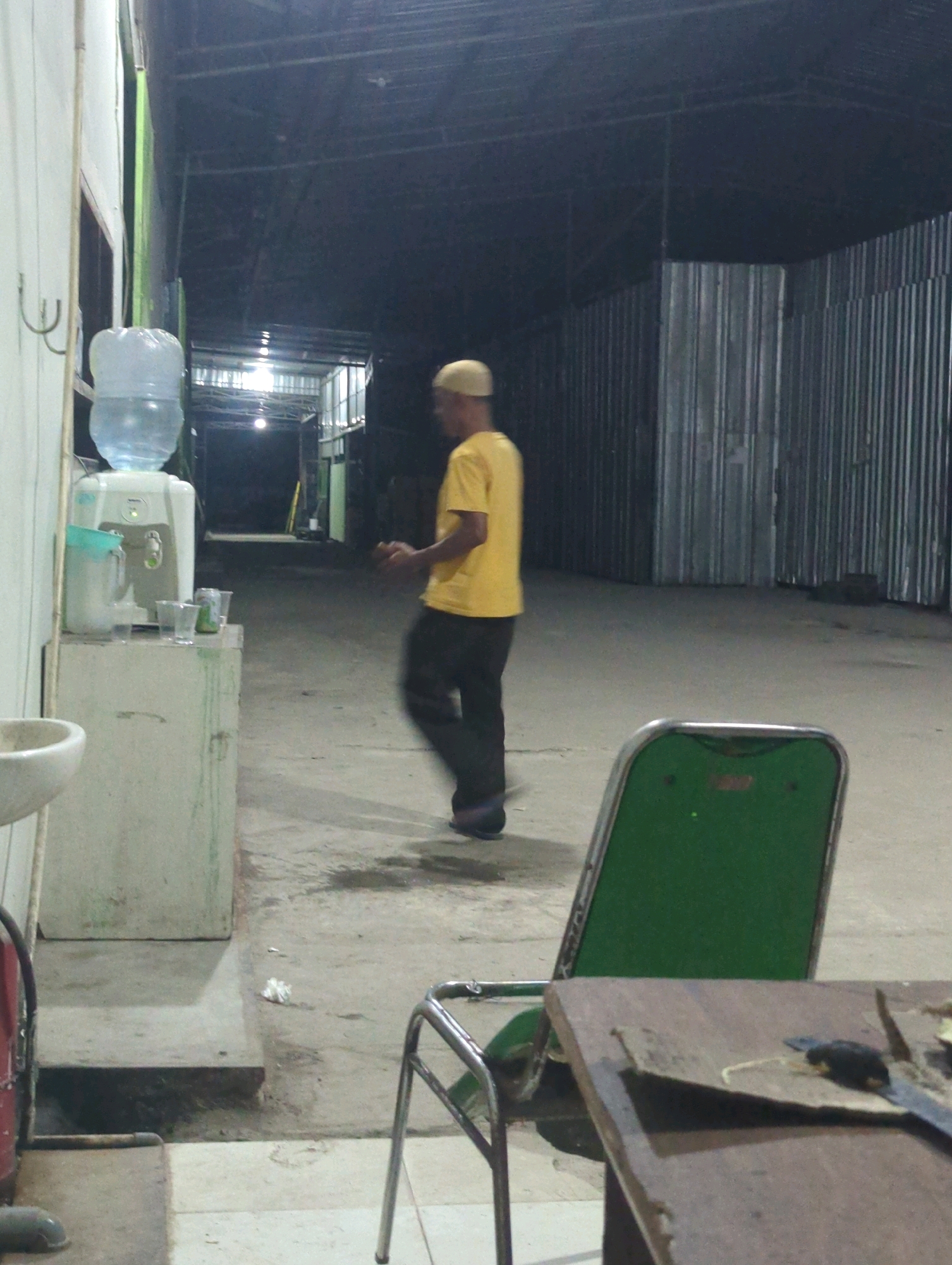 The image depicts an outdoor, covered area with a concrete floor, likely a workshop or storage space, located in Batu Belaman, Indonesia. A man wearing a yellow t-shirt and a beige cap is in motion, possibly walking or dancing, in the middle ground. To the left, a white water cooler with a large blue water bottle on top is situated on a light-colored cabinet. Several clear plastic cups and a can are placed on the cabinet next to the water cooler. A white sink basin is visible to the left of the cabinet. In the foreground, a green chair with a metal frame is positioned near a wooden table, which has some objects on its surface. The environment appears somewhat unkempt, with a corrugated metal wall forming the background on the right side of the image. The lighting suggests it is either evening or indoors with artificial illumination.