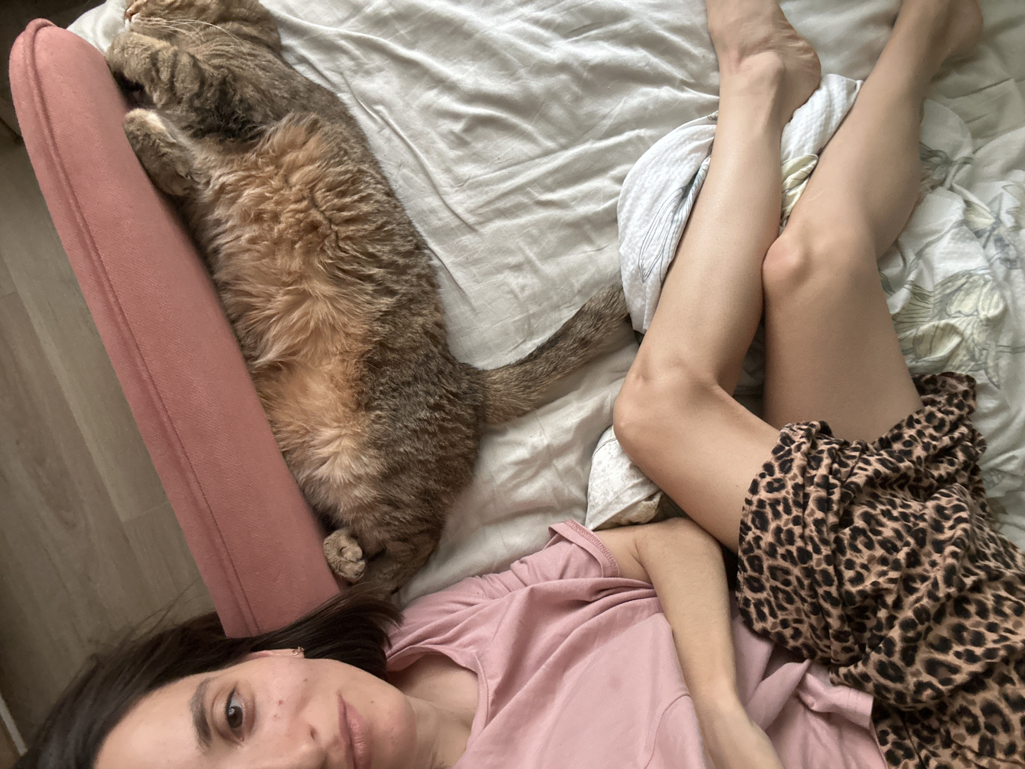 An overhead shot captures a woman and a cat relaxing together indoors, likely in a bedroom in Moscow, Russia. The woman lies on her back, looking directly at the camera, seemingly taking a selfie. She has dark hair, dark eyes, and a neutral, relaxed expression. She is wearing a plain pink t-shirt, and a brown and black leopard-print garment or blanket covers her lower body and legs, which are partially bent.
Next to her, a fluffy tabby cat with brown, gray, and black fur is stretched out, also lying on its back, its paws tucked near its head. The cat is positioned alongside a salmon-pink upholstered cushion or armrest. Both the woman and the cat are on a bed or a soft surface covered with light-colored, possibly off-white or beige, wrinkled bed linens. A small white pillow with a subtle pattern is visible near the woman's legs. The lighting suggests it is daytime, with soft natural light illuminating the scene. No visible text is present in the image.