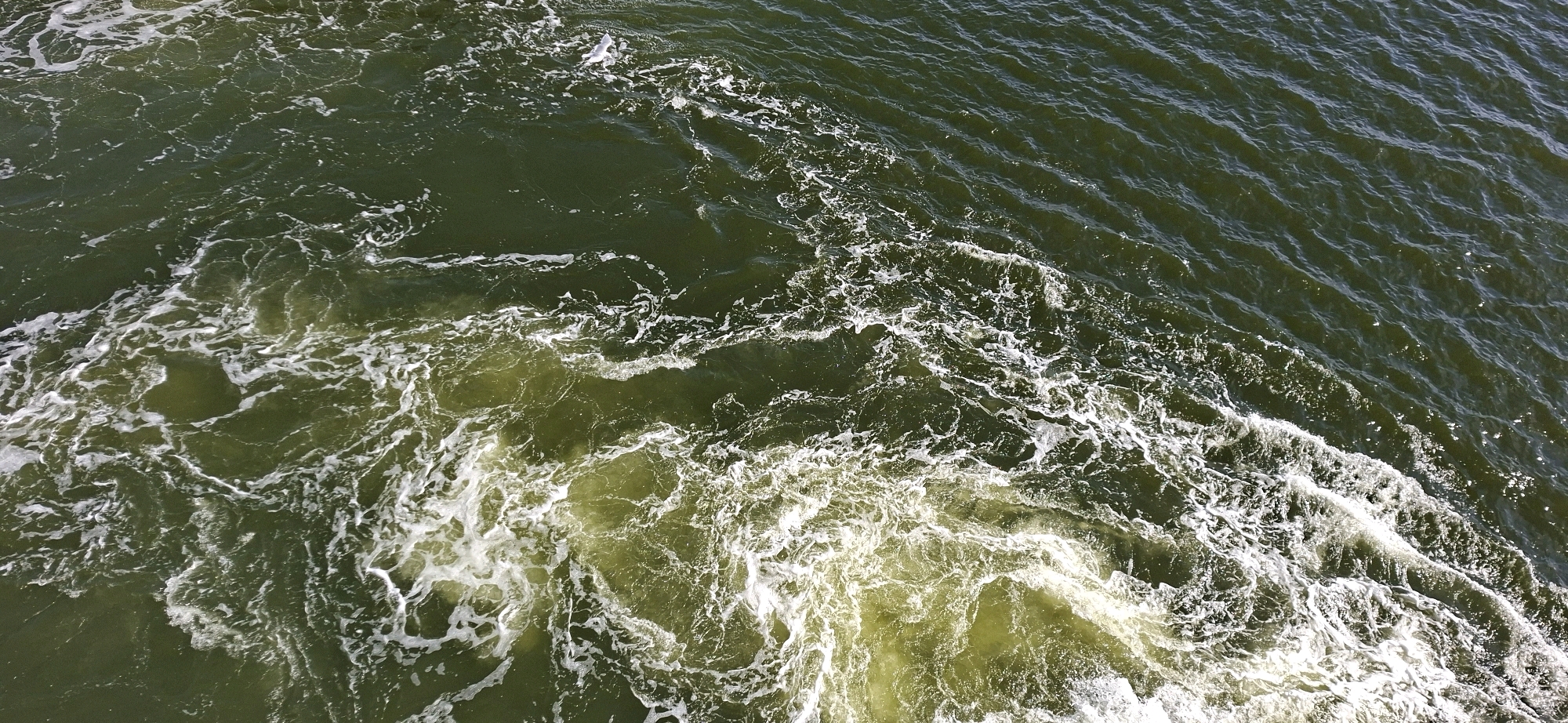 This is an aerial, high-angle, medium shot of rough, foamy green water in a body of water. The water is agitated, with white foam and swirling patterns indicating movement. The surface of the water has small ripples that create a textured effect. The lighting suggests it is daytime. There are no visible people, animals, or man-made objects in the frame. The scene depicts a dynamic water surface, potentially a wake from a vessel or a natural disturbance. Without additional context, the specific location in Helsinki, Finland, cannot be determined, but it could be a harbor, the sea, or a large lake within the city.