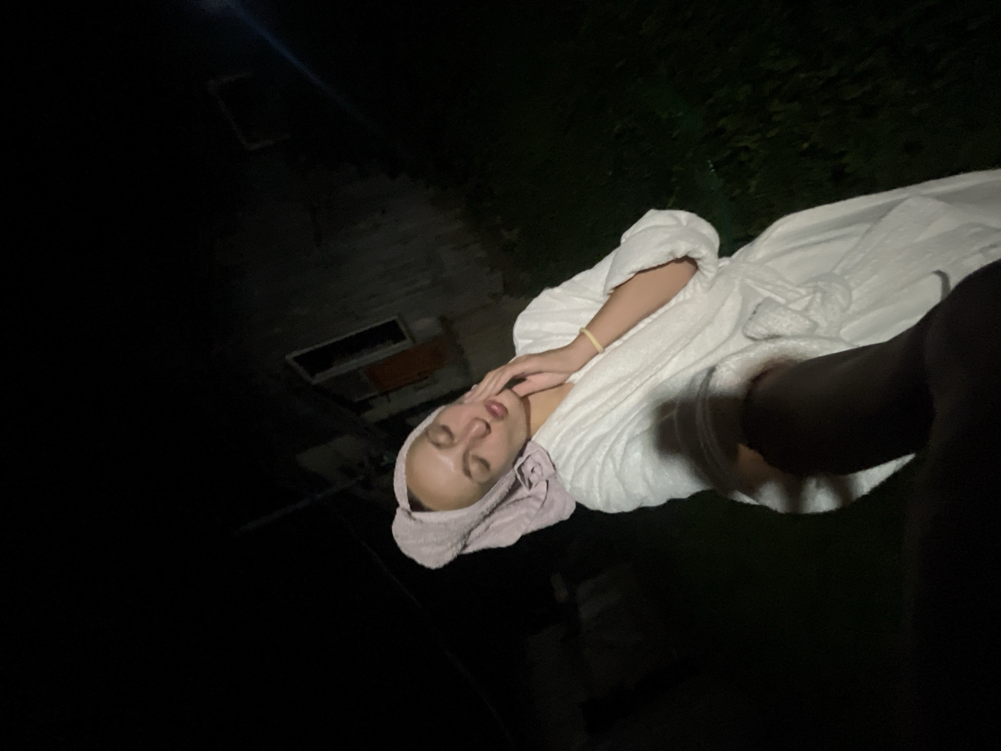 A young woman, wearing a white bathrobe and a towel wrapped around her head, stands in an outdoor, dimly lit environment at night. She is captured in a selfie-style shot, with her left hand raised towards her face, touching her cheek. Her eyes are closed, and her lips are pursed. A hint of a yellow bracelet is visible on her left wrist. Behind her, a dark, dilapidated structure, possibly a shed or an old house, is faintly visible against the night sky. The ground in front of the structure appears to be covered in grass. The image has a slightly grainy quality, typical of low-light photography. The location is Sharkan, Russia.