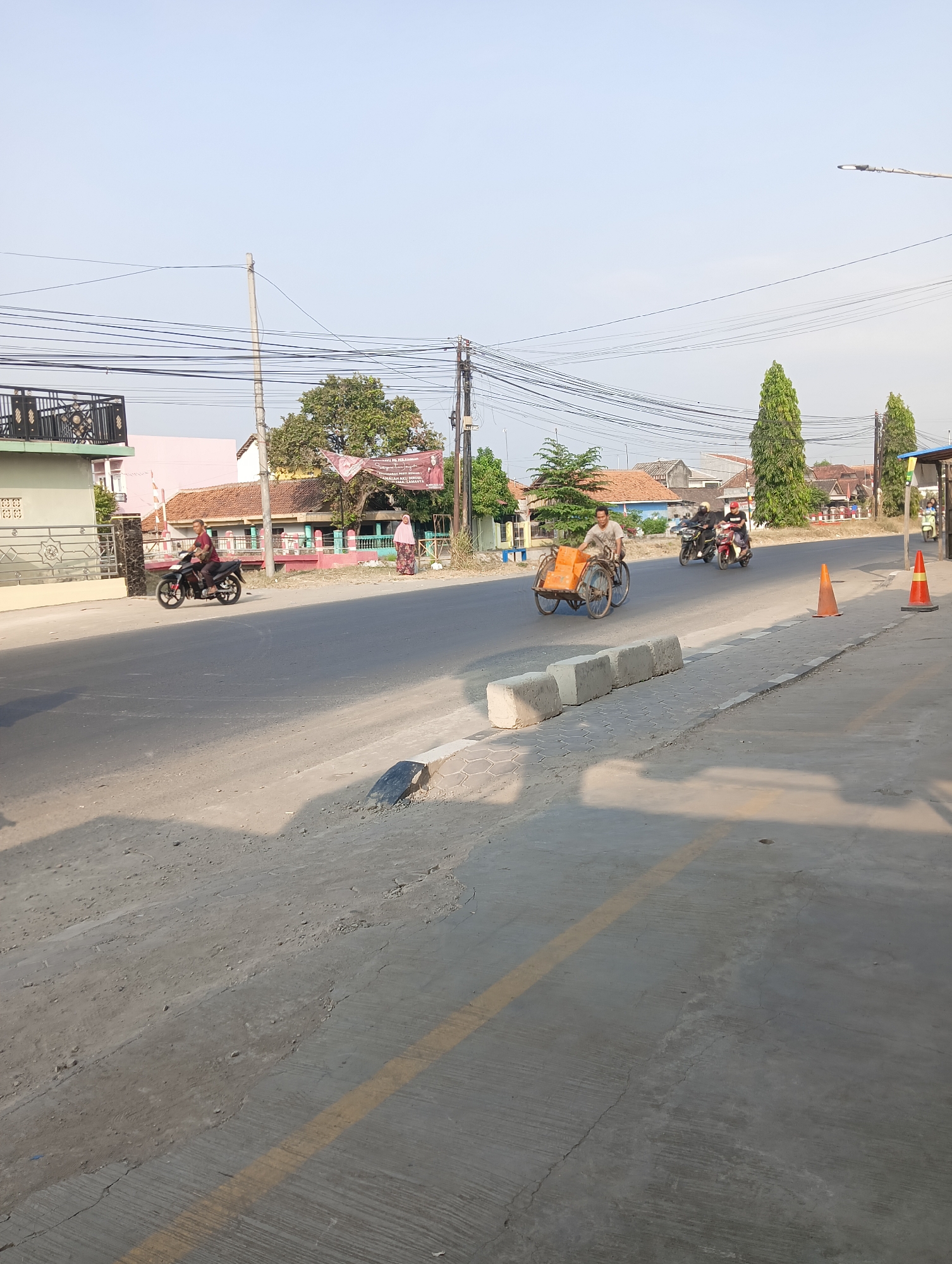 The image depicts a street scene in Leuweunggajah, Indonesia. The foreground is a concrete road with a yellow line painted on it. Several concrete blocks are placed along the side of the road, which is lined with trees and buildings. On the road, a man is riding a motorcycle, a motorized rickshaw carrying boxes is being pedaled by a man, and two other motorcycles are visible in the distance. A woman in a headscarf is standing on the side of the road. Power lines are strung across the sky. The sky is clear and blue.