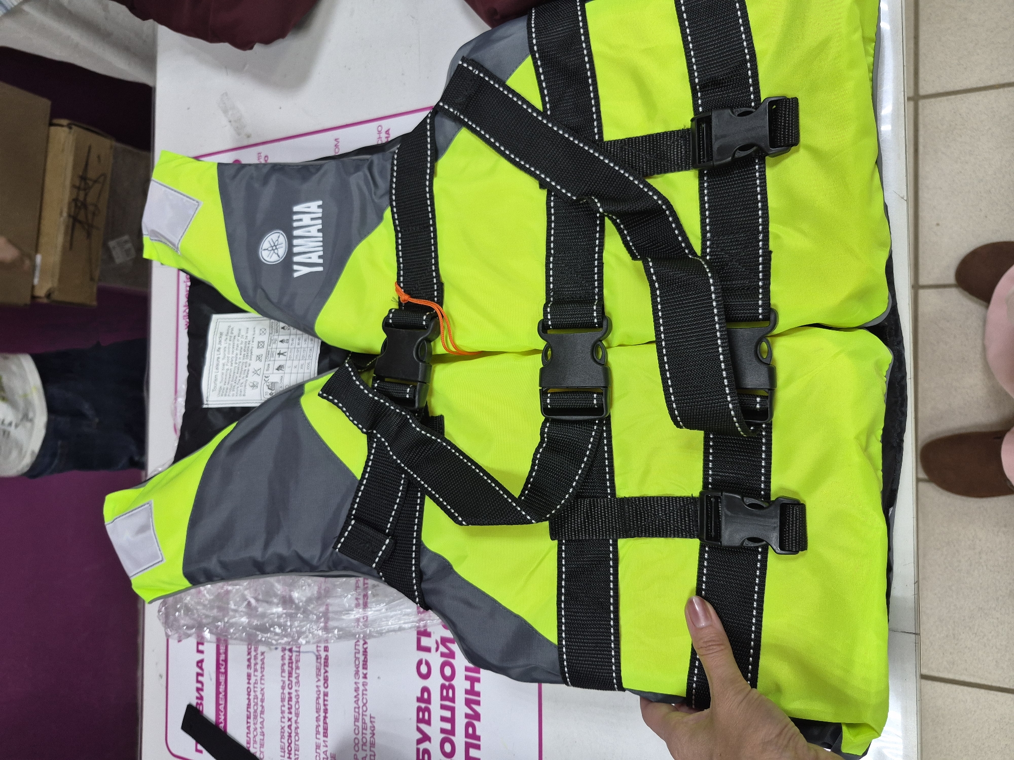 The image shows a bright yellow and gray Yamaha life jacket laid flat on a white surface. The life jacket has multiple black straps with buckles for securing it. A person's hand, with painted fingernails, is visible on the left side of the image, holding the life jacket. In the background, there is a white paper with text in Cyrillic alphabet and a clear plastic bag. The overall environment appears to be indoors, possibly a retail or storage space. There is no identifiable information or context to place this image specifically within the city of Chaikovsky, Russia.