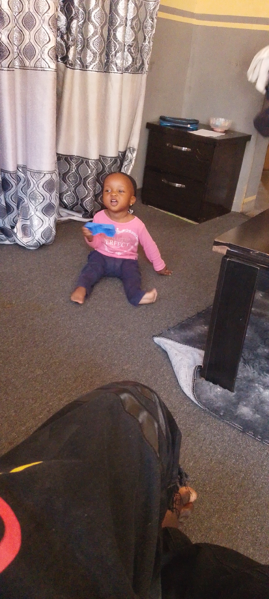 A young child, appearing to be a toddler, is sitting on a carpeted floor in a room. The child is wearing a pink long-sleeved shirt with a heart design and the word "PERFECT" on it, along with dark blue pants. The child is holding a blue object in their right hand and looking to the left of the frame. The background includes a dark dresser with silver drawer pulls, draped curtains with a patterned design, and a portion of a dark table. The floor appears to be a dark gray carpet. The image also shows the legs and feet of an adult in the foreground, wearing dark clothing. The scene suggests an indoor domestic setting, potentially in a residential area of Dorowa, Nigeria.