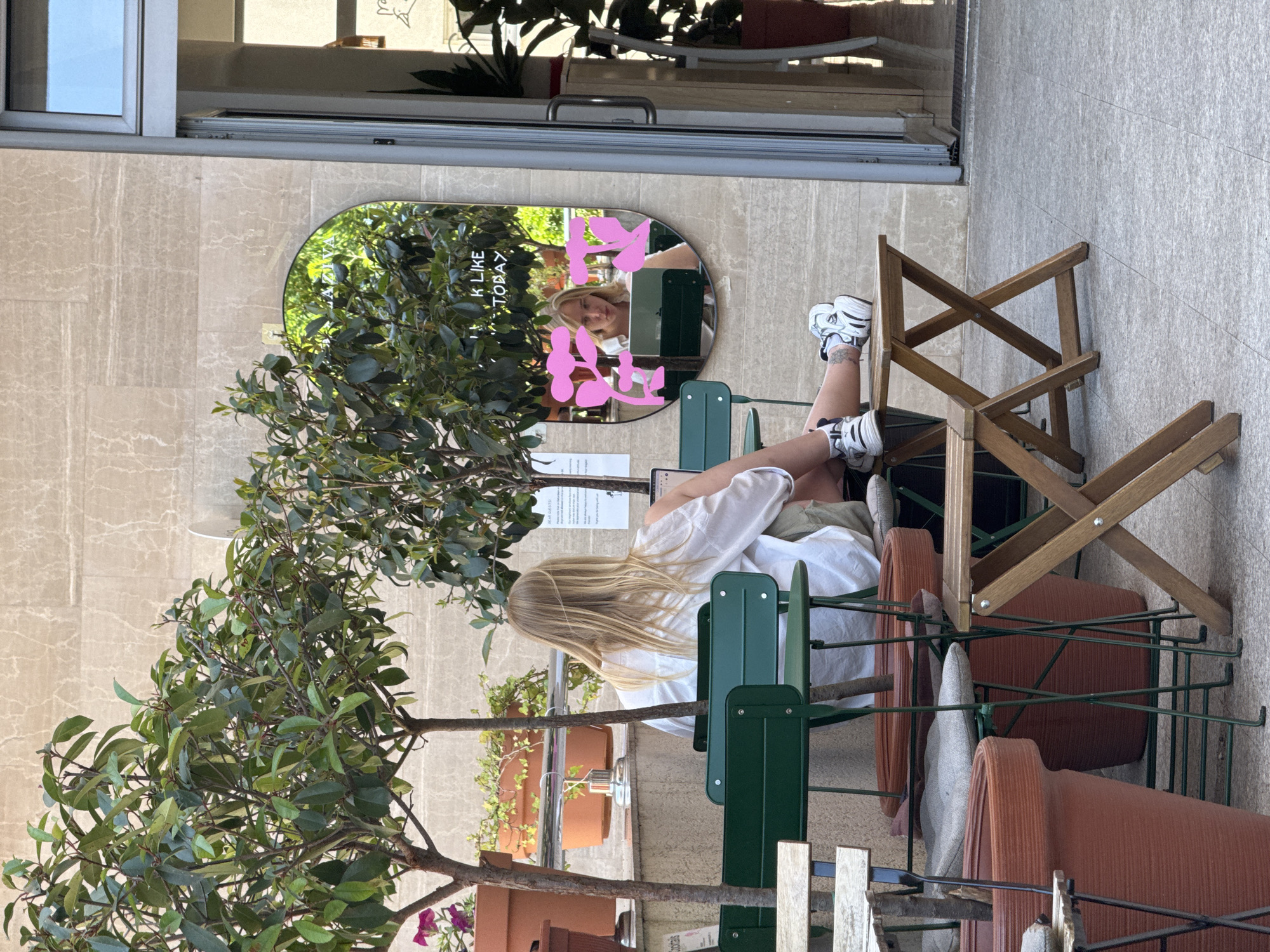 A woman with long blonde hair sits at an outdoor cafe in what is likely Budva, Montenegro. She wears a white shirt and sneakers, her legs crossed, one foot propped on a green metal chair. A leafy tree is beside her, along with stacked clay pots containing plants. Behind her, a mirror reflects her face with pink graffiti-like designs. Another chair, made of wood, is close by. The cafe appears to be next to a building with a glass-paned door.
