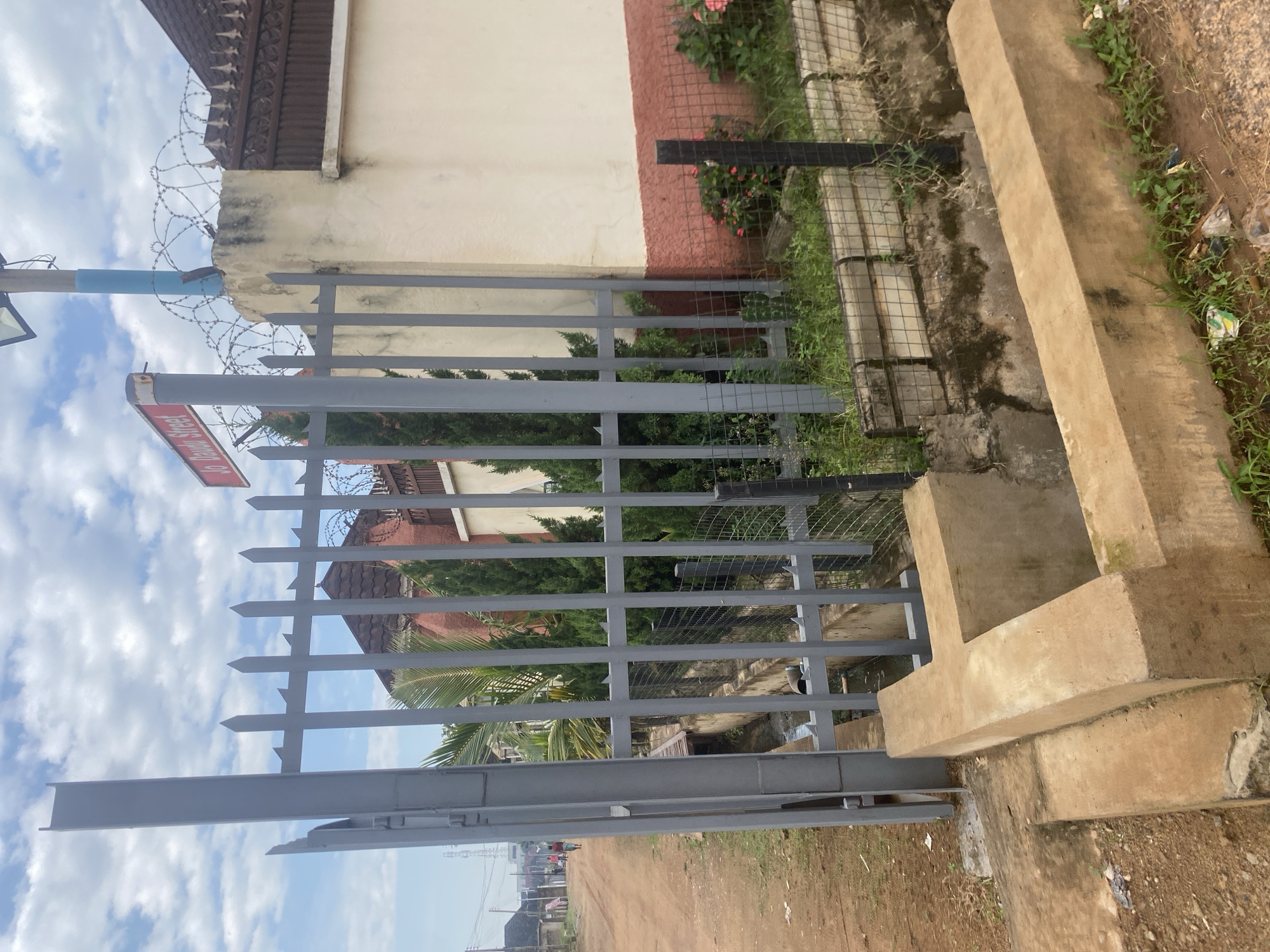 The image shows a view of a street entrance in Barden Yana, Nigeria. A gray metal gate stands open, revealing a concrete drainage system in front of it. Beyond the gate, a dirt road stretches into the distance, with a few people walking along it. On the right side of the image, a white and red wall is topped with barbed wire, and a street sign labeled "Jo Daudu Street" is visible. There are trees and bushes planted behind the fence. The sky is cloudy.