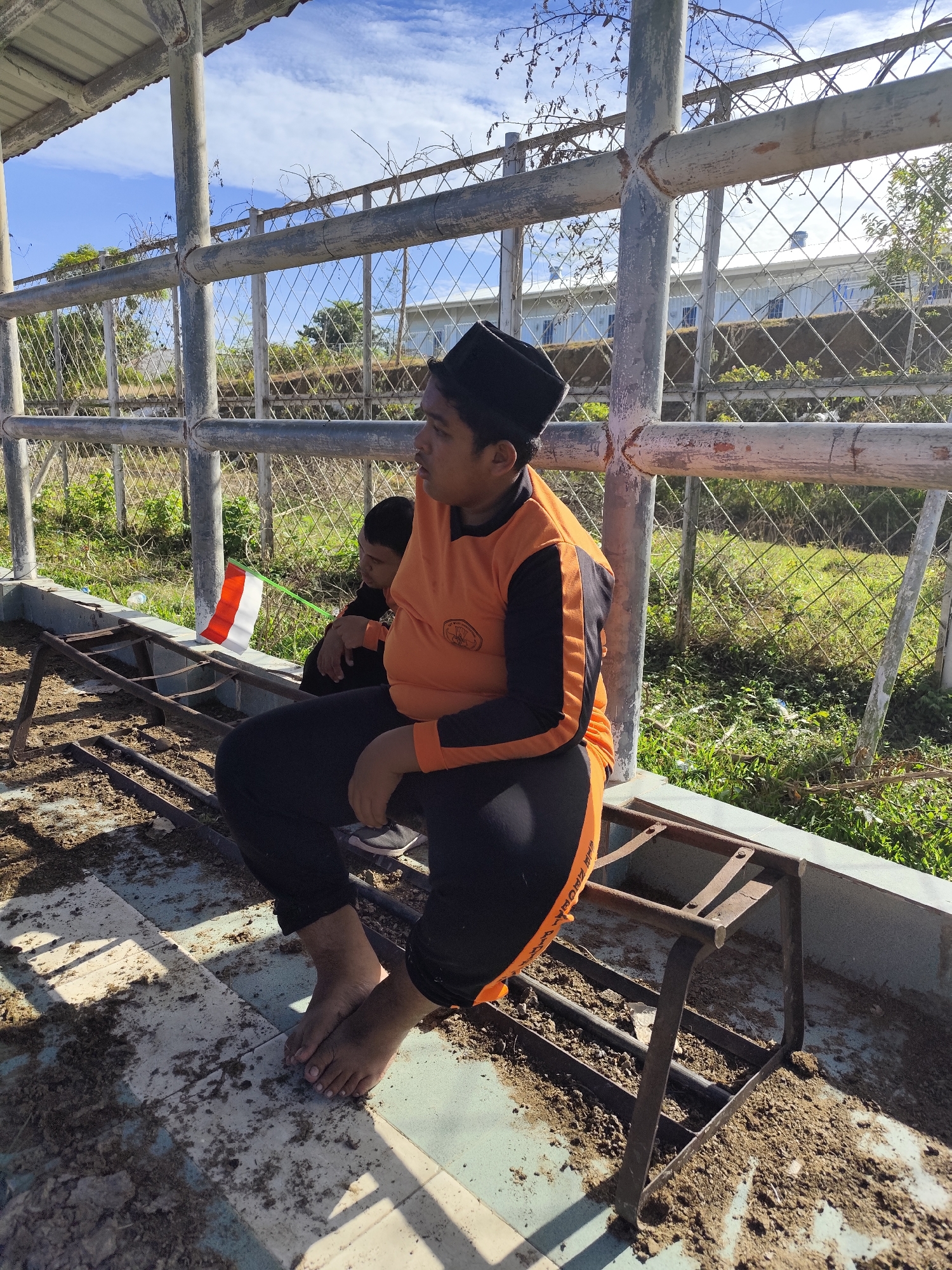 An individual wearing an orange and black long-sleeved shirt, black pants, and a black cap sits on a metal bench, facing right, with their bare feet visible. To their left, another individual, partially obscured, sits behind them. A small Indonesian flag is placed on the bench between the two individuals. The scene appears to be outdoors, possibly under a covered structure with metal supports. The ground is dirty and covered with debris. In the background, a fence separates the area from a grassy field and a building. The sky is blue with some clouds.