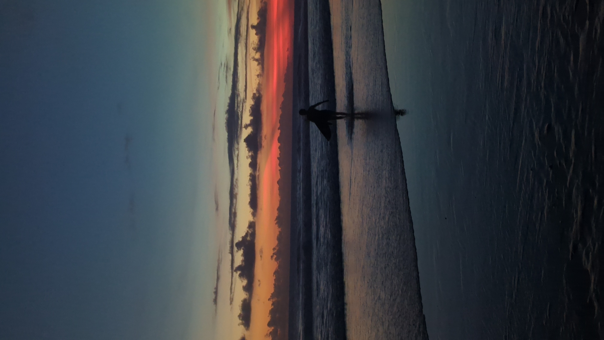 The image depicts a surfer walking on a beach at sunset in North Kuta, Indonesia. The sky is divided into a deep blue on the left and vibrant hues of orange and red where the sun is setting. Dark, vertical clouds punctuate the sunset's gradient. The surfer, a dark silhouette, is positioned in the middle of the frame, holding what appears to be a surfboard. The beach is a dark, wet sand with subtle ripples from the receding waves. The water's edge reflects the colors of the sky, creating a mirrored effect.