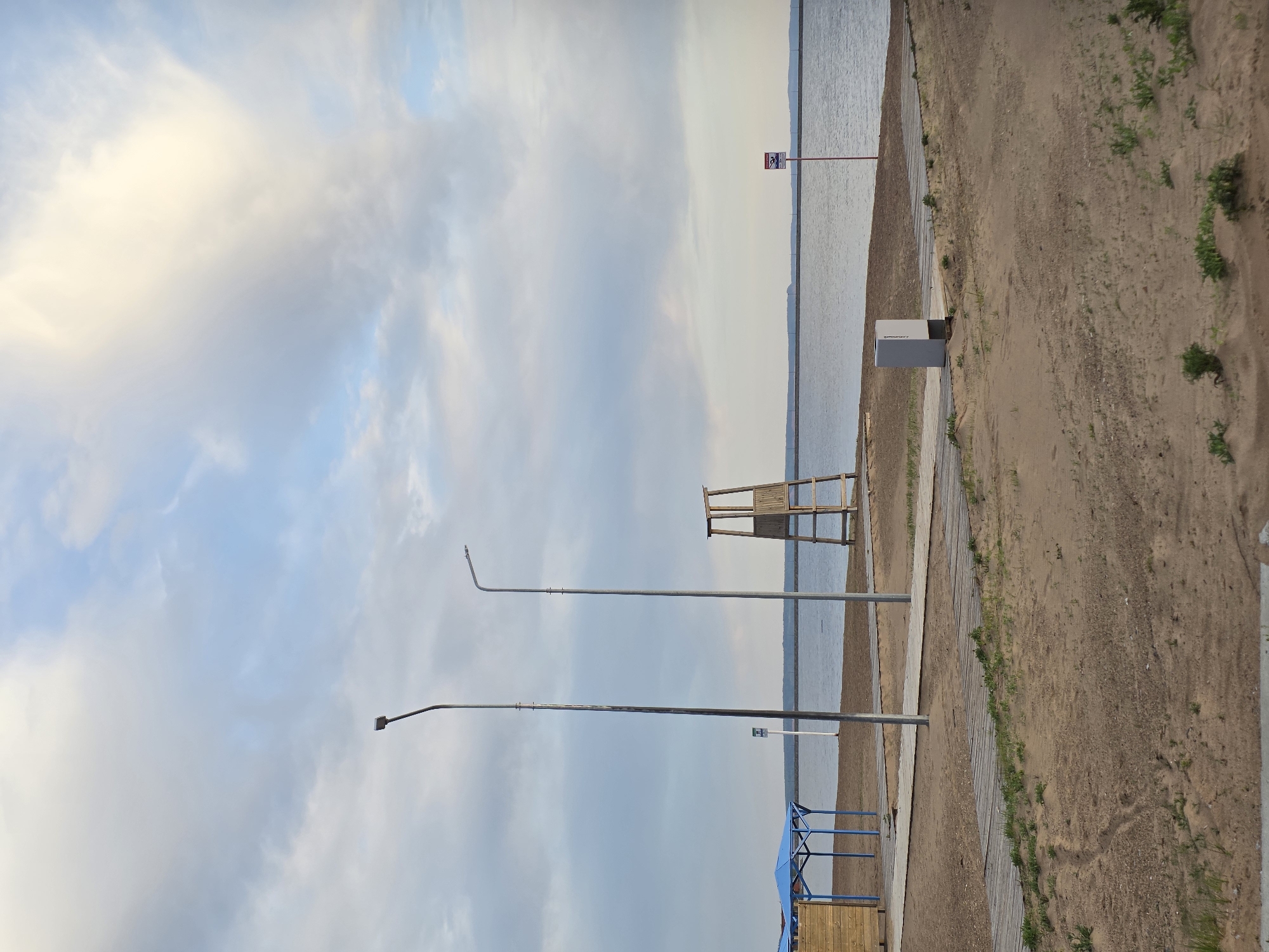 The image shows a beach scene in the city of Chaikovsky, Russia. The scene includes a sandy beach, a body of water (likely a lake or river), and a cloudy sky.
Several objects are visible on the beach, including:
* A lifeguard chair
* A wooden gazebo or shelter with a blue roof
* Two tall streetlights
* A small gray utility box
* A sign on a pole near the water's edge
The beach appears to be mostly empty, with no people visible. The environment is relatively natural, with some vegetation growing along the edge of the beach.