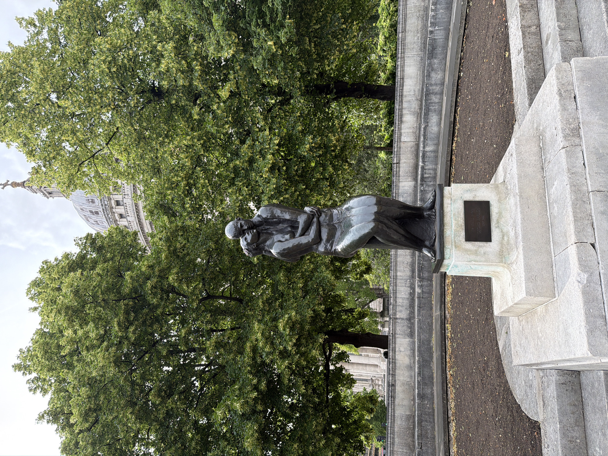 This image captures a bronze sculpture of two embracing figures in a public space, identified as the gardens near St Paul's Cathedral in London, United Kingdom.
The central subject is a dark bronze statue depicting a male and female figure in a standing embrace. The female figure is positioned to the viewer's left, leaning into the male figure on the right, with their arms interlocked around each other's backs. The statue rests on a multi-tiered pedestal composed of light grey, rough-hewn stone blocks. A dark, rectangular plaque is affixed to the lower front face of this pedestal.
Immediately surrounding the pedestal, a patch of dark, bare soil is visible, containing a sparse scattering of dry, brown leaves. This area is enclosed by a low, curved retaining wall and a series of wide, shallow steps made of the same light grey stone, extending across the foreground and midground.
In the midground and background, two large deciduous trees with dense green foliage frame the scene. The tree on the left features a thick trunk and extends upwards, partially obscuring a building with visible columns to its left. The tree on the right also has extensive green leaves, and behind its upper canopy, the iconic grey dome, lantern, and cross finial of St Paul's Cathedral are clearly discernible against a bright, overcast sky. The overall lighting is soft and diffused, consistent with a cloudy day.