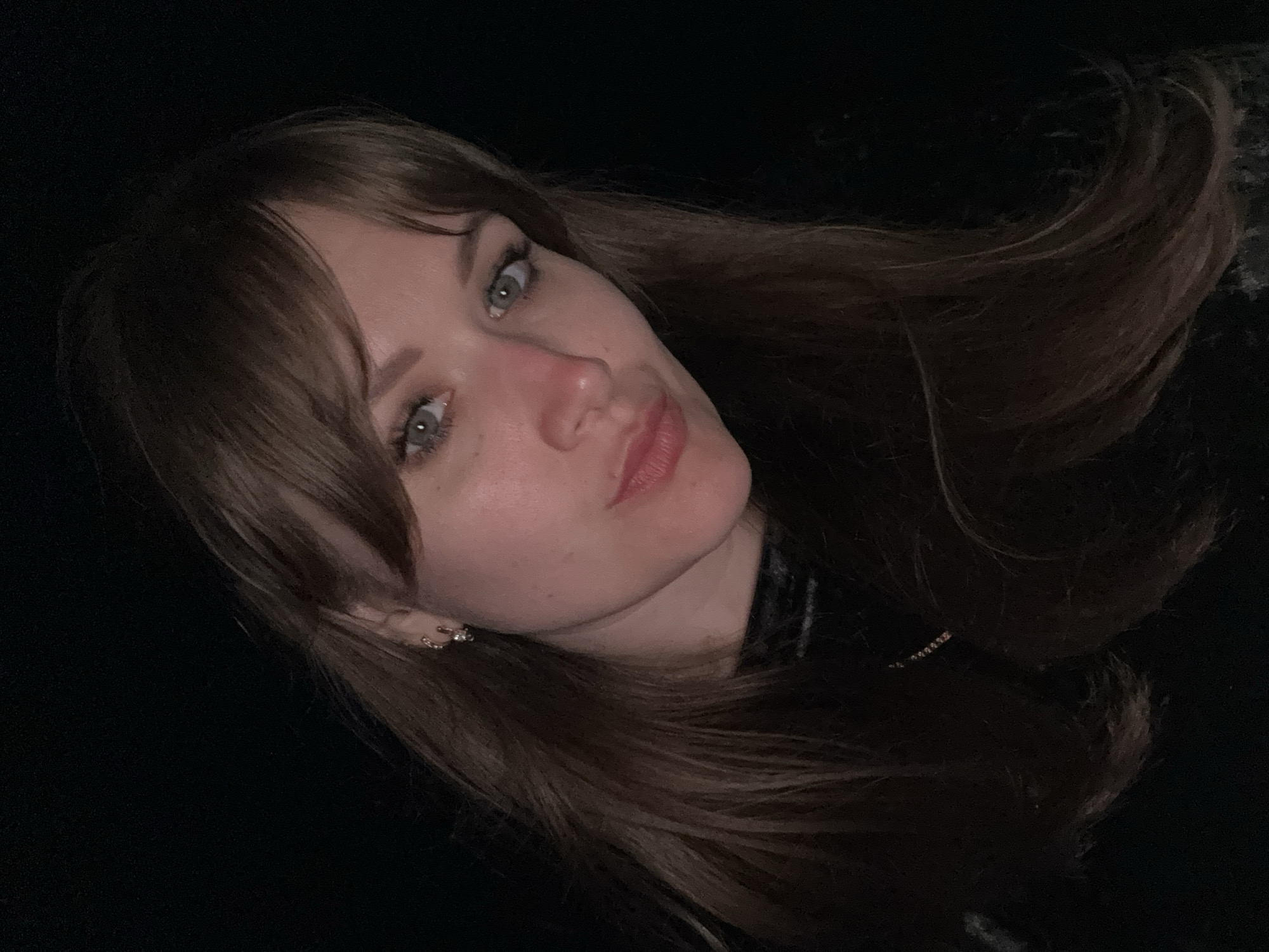 A photograph of a woman with light skin and long brown hair. She has bangs and her hair is styled loosely. She is wearing an earring on her left ear. The background is mostly dark, suggesting a low-light environment. There is no information about her location.