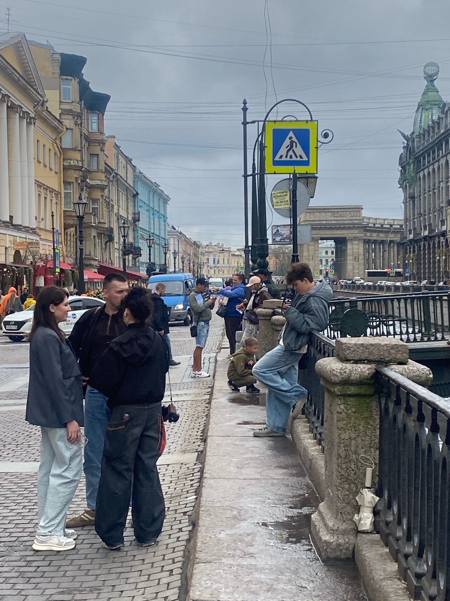 The image depicts a street scene in Saint Petersburg, Russia, likely along a canal. In the foreground, a group of people are gathered on a cobblestone sidewalk adjacent to a railing overlooking a body of water. A woman in a dark blazer and light-colored pants stands with her side to the camera, engaged in conversation with two men. One man, wearing a black jacket and blue jeans, faces the woman, while another man in a black hooded jacket stands between them, looking away. To the right, a young person in a grey hooded jacket and baggy blue jeans leans against the railing, looking at their phone. Further down the sidewalk, a small child in a green jacket crouches. Across the street, a blue van and other vehicles are visible, along with a row of multi-story buildings in various architectural styles and colors. A pedestrian crossing sign with a blue triangle and white pedestrian symbol is mounted on a lamppost. The sky is overcast, contributing to a muted, somewhat somber atmosphere.