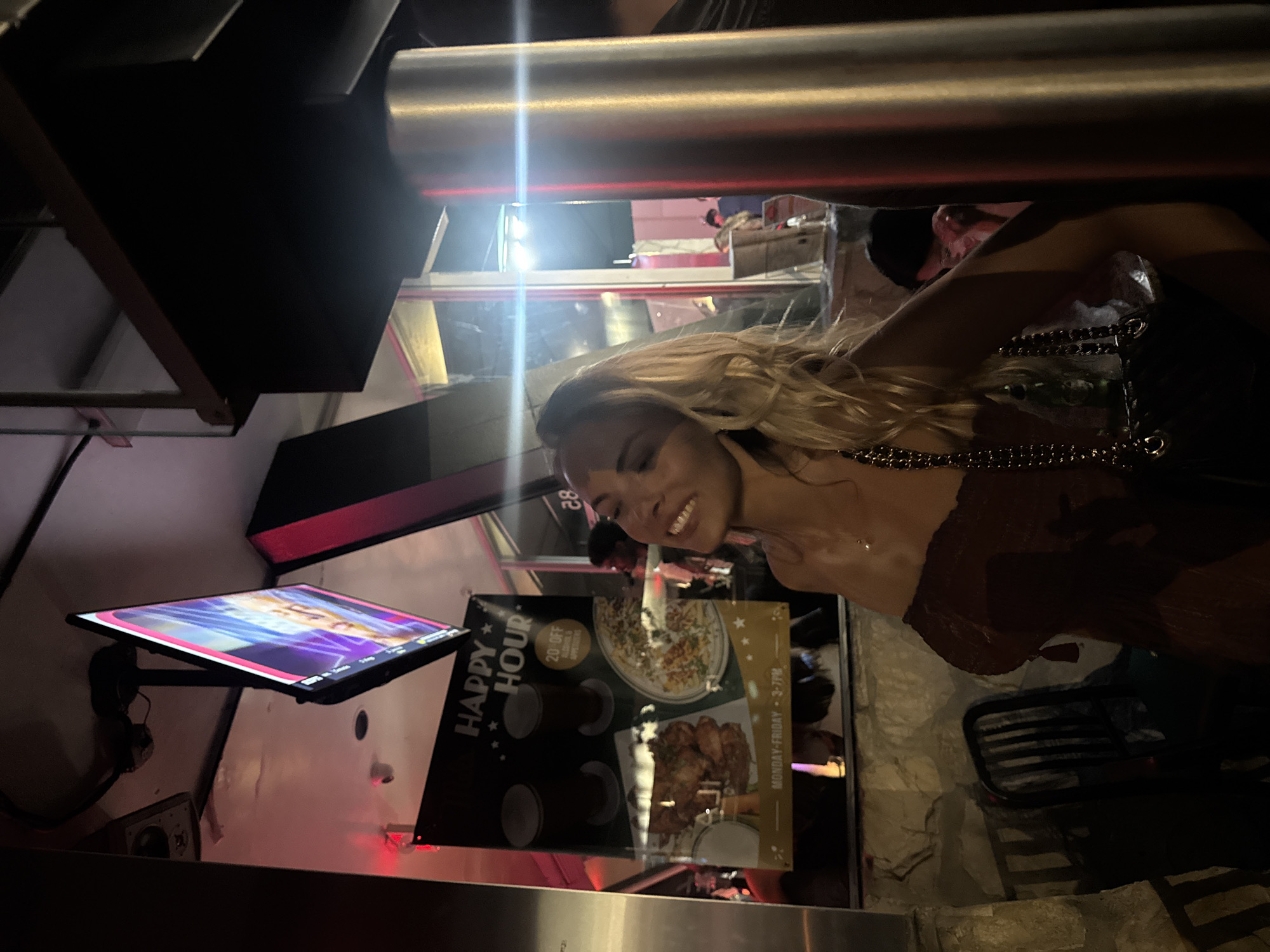 The image shows a woman with blonde hair smiling in what appears to be a bar or restaurant. The restaurant has a happy hour promotion for 20% off alcohol and appetizers. The restaurant is open for happy hour on Monday - Friday from 3-7PM. The location of the restaurant is in West Hollywood, United States of America.
