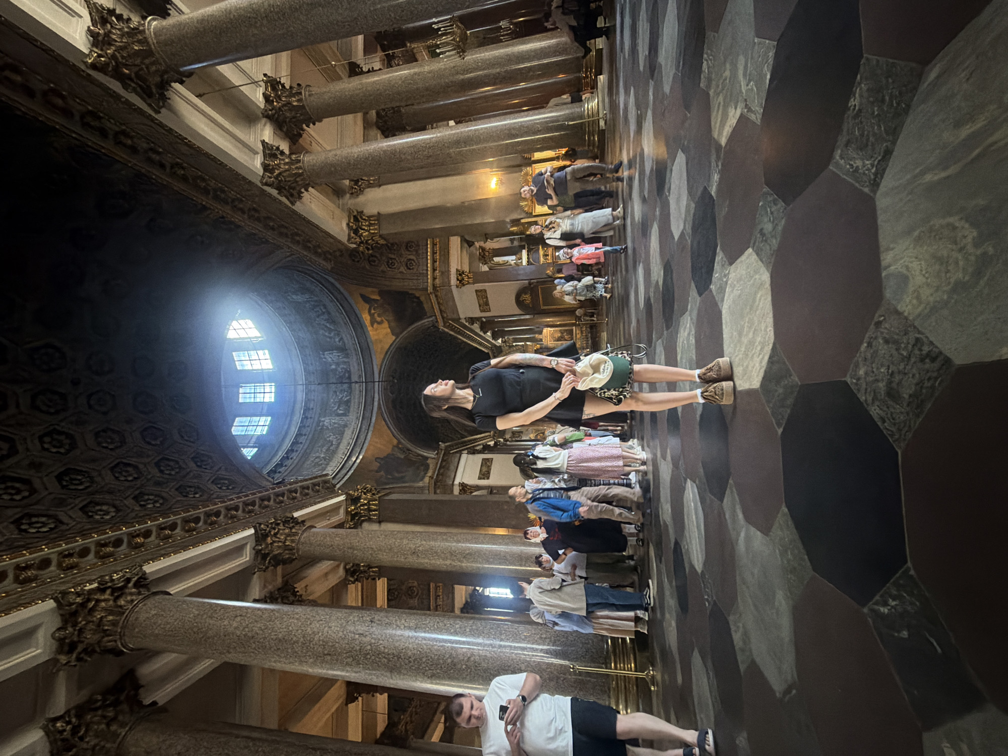 An interior shot captures the grandeur of a cathedral or large domed building, emphasizing its opulent architecture. The high, vaulted ceiling features intricate ornamentation and a central skylight bathing the space in natural light. Massive columns, some with elaborate gold capitals, punctuate the vast hall, casting shadows on the patterned stone floor. In the foreground, a young woman stands, looking up and to the right, holding a light-colored hat. She is dressed in a black short-sleeved top and a black skirt, with casual shoes. To her left, a man in a white t-shirt and black shorts is engrossed in his phone. Behind them, a diverse group of people, likely tourists, are scattered throughout the hall, some engaged in conversation, others observing the surroundings. The flooring is a striking mosaic of large, geometric stone tiles in shades of gray, black, and brown, creating a visually dynamic surface. The overall impression is one of awe and historical significance, with the architectural elements dominating the scene.