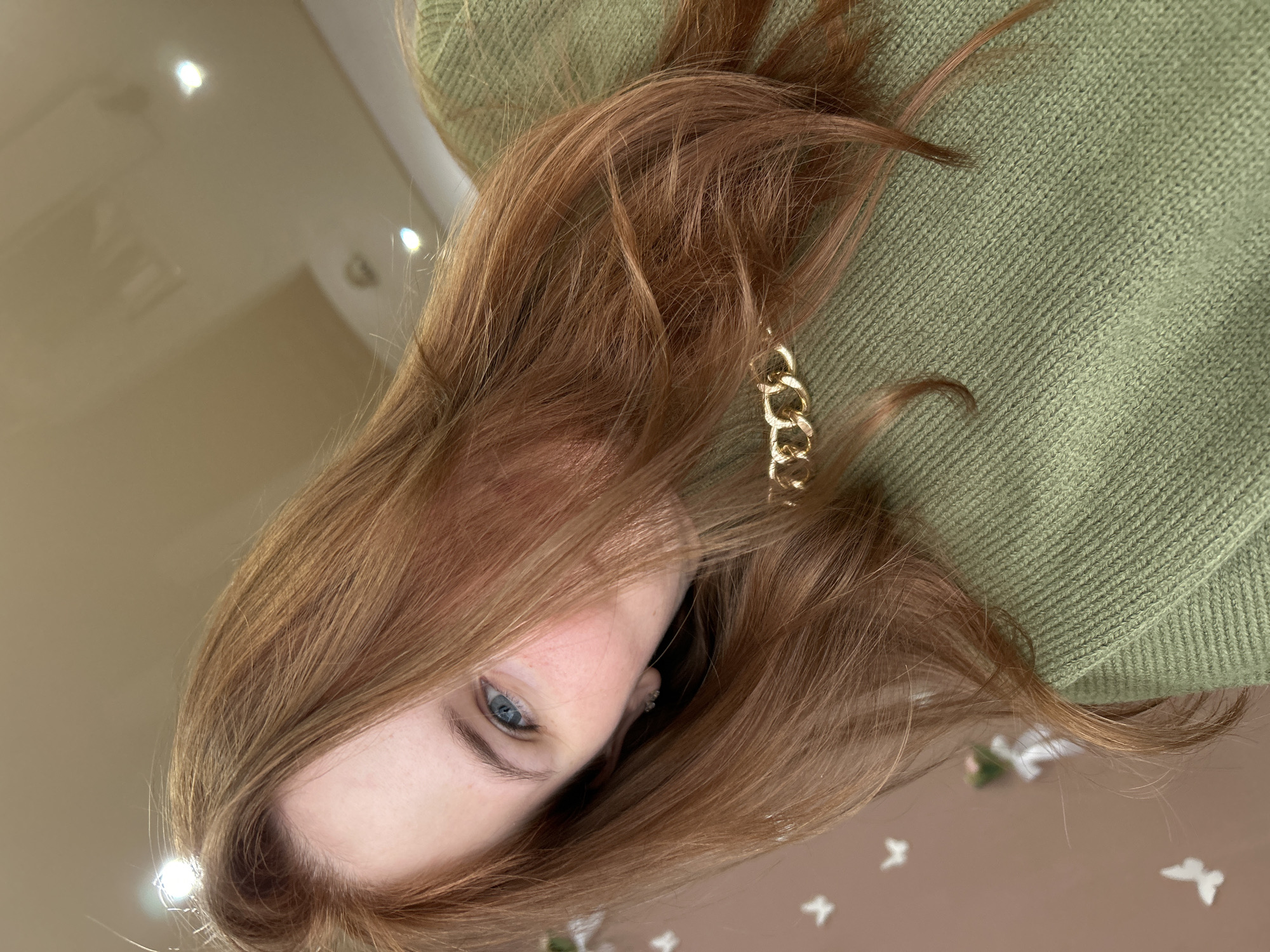 A tilted, close-up perspective features a person with light brown hair, blue eyes, and fair skin. They are wearing a green sweater and a gold chain earring is visible. The background appears to be a beige or light brown surface with subtle patterns or lighting effects. There is no information to infer that this image was taken in Chaikovsky, Russia.